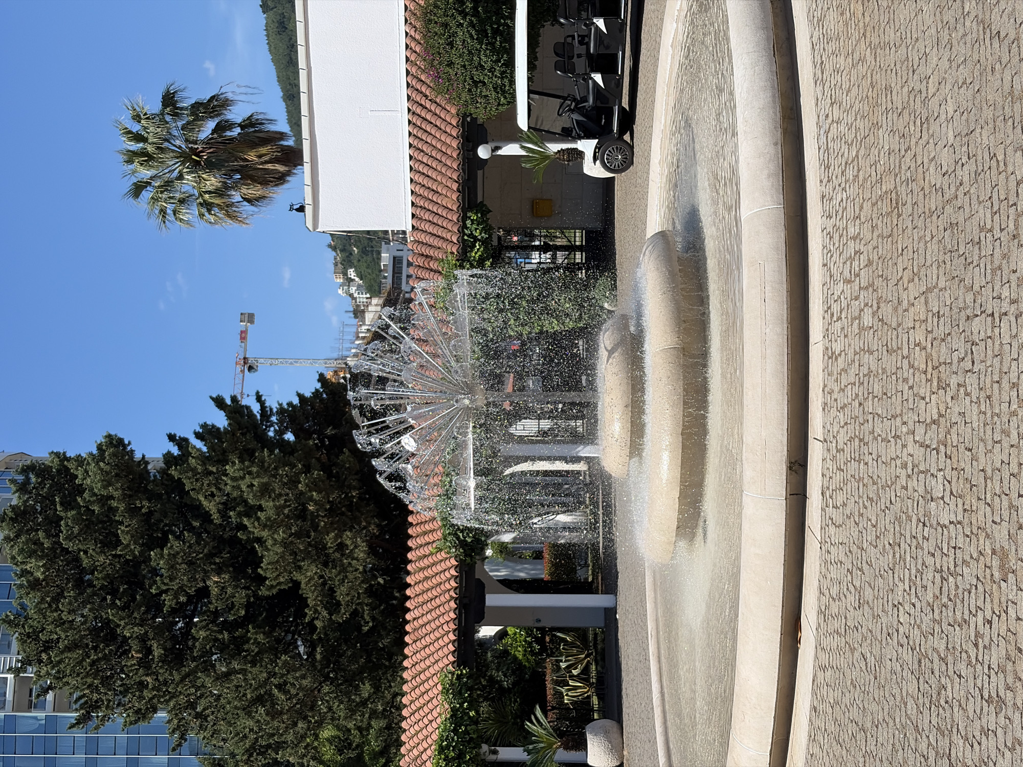 The image shows an outdoor scene in Budva, Montenegro, featuring a tiered fountain with a central spraying mechanism. The fountain is constructed of light-colored stone and surrounded by a paved area. Adjacent to the fountain is a building with a red-tiled roof and white exterior. A golf cart is parked next to the building. Trees and greenery are visible behind the building and fountain. A palm tree is also present, set against a clear blue sky. A construction crane can be seen in the background. The scene appears to be well-maintained and located in a tourist or resort area.