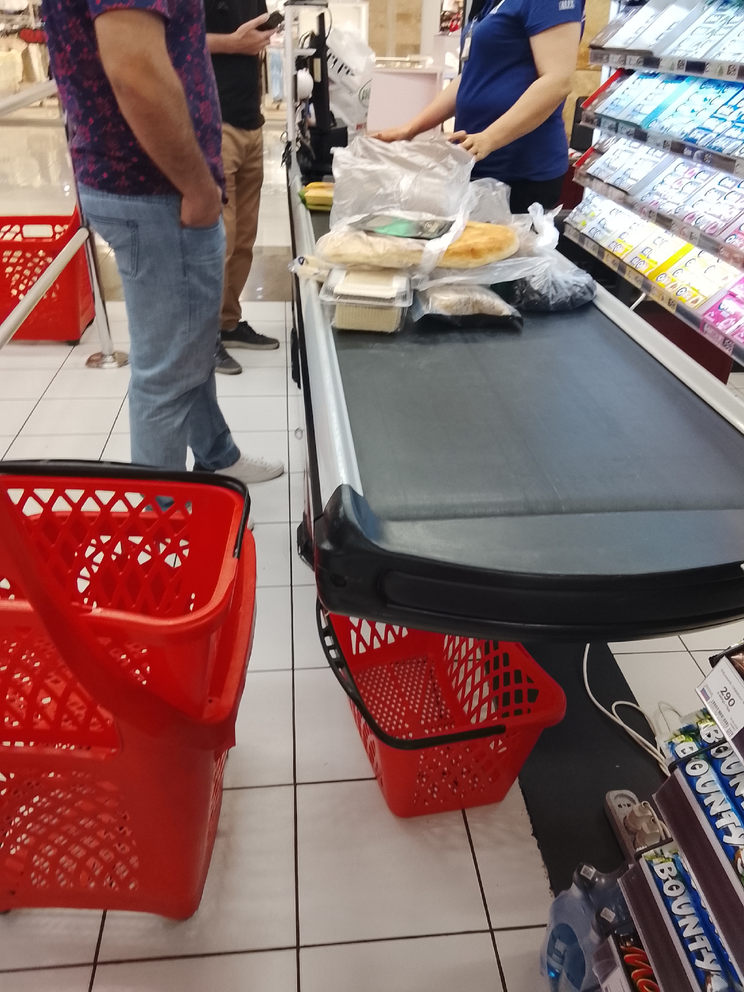 The image shows a close-up view of a checkout counter at a grocery store. In the foreground, two red plastic shopping baskets are visible. The left basket is mostly empty, while the right basket contains several items, including packaged goods and what appears to be bread. Behind the baskets, a conveyor belt runs across the frame, with various grocery items placed on it. To the left of the counter, a person wearing a blue shirt and black pants is standing, their hands resting on the conveyor belt. To the left of this person, another person wearing a patterned shirt and jeans is standing. Further to the left, part of a checkout machine and a display of packaged goods can be seen. The floor is tiled in a grid pattern. The overall lighting suggests an indoor retail environment.