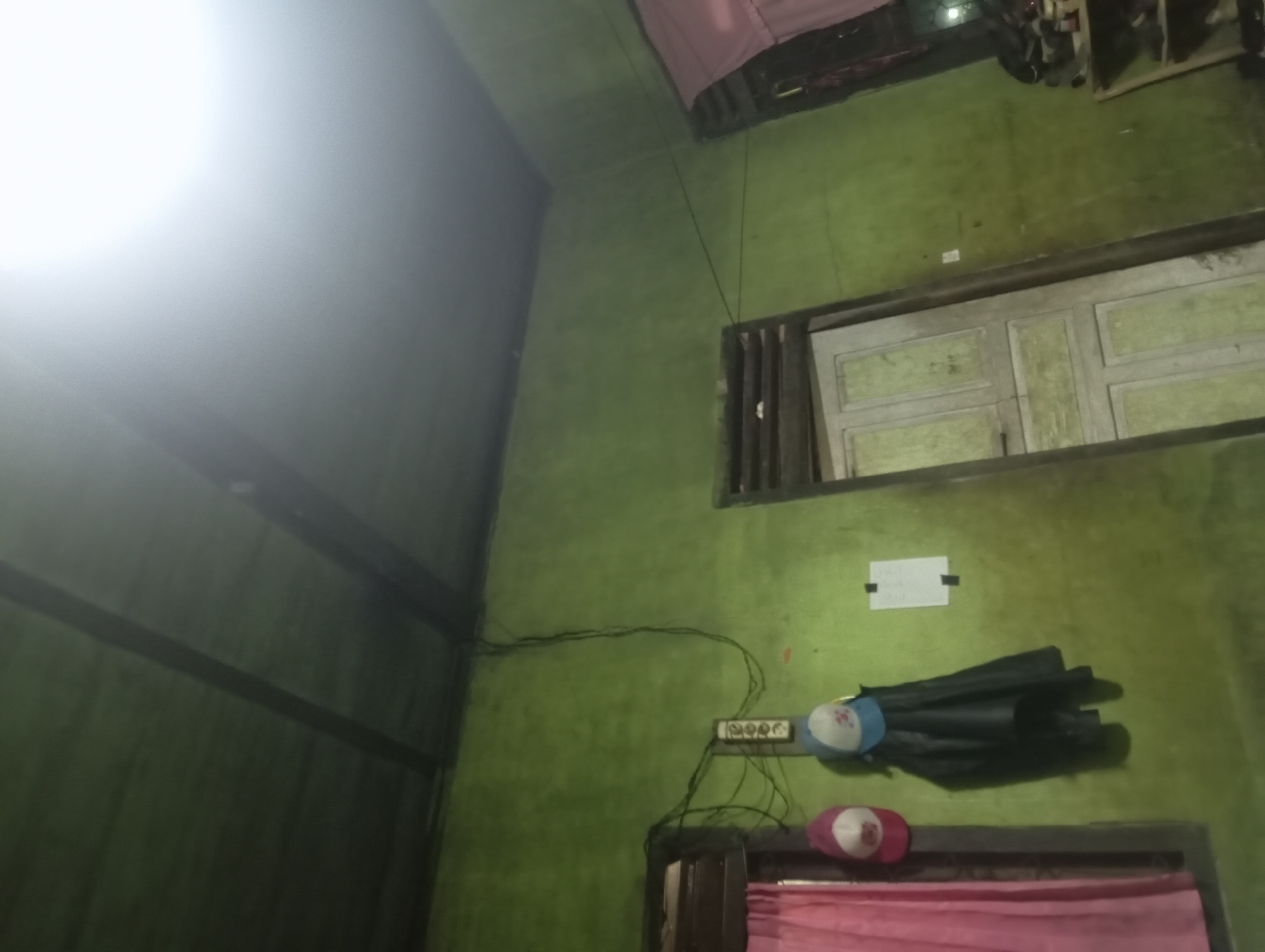 The image depicts an interior view of a room, likely in a residential setting in Rejosari, Indonesia. The walls are painted a vibrant green, and the ceiling appears to be made of dark wood or metal panels with visible beams. A bright light source, possibly a ceiling light or sunlight, is intensely illuminating the upper right portion of the frame, creating a blown-out effect.
In the foreground, on the left side of the image, is a window with a pink curtain. Below the window, a dark-colored jacket is hanging, and two caps are placed on a hook or nail. One cap is red and white, and the other is blue and white. A vertical electrical box with multiple outlets is mounted on the wall between the window and the caps. Several black electrical wires are visible, running along the wall and ceiling.
To the right of the caps, a white piece of paper is affixed to the wall with black tape. Further to the right, a wooden door with multiple panels and a distressed finish is visible. The door is slightly ajar. On the far right edge of the image, a shelf unit is partially visible, with several shoes and other items on its shelves. The overall impression is of a somewhat cluttered, utilitarian living space.