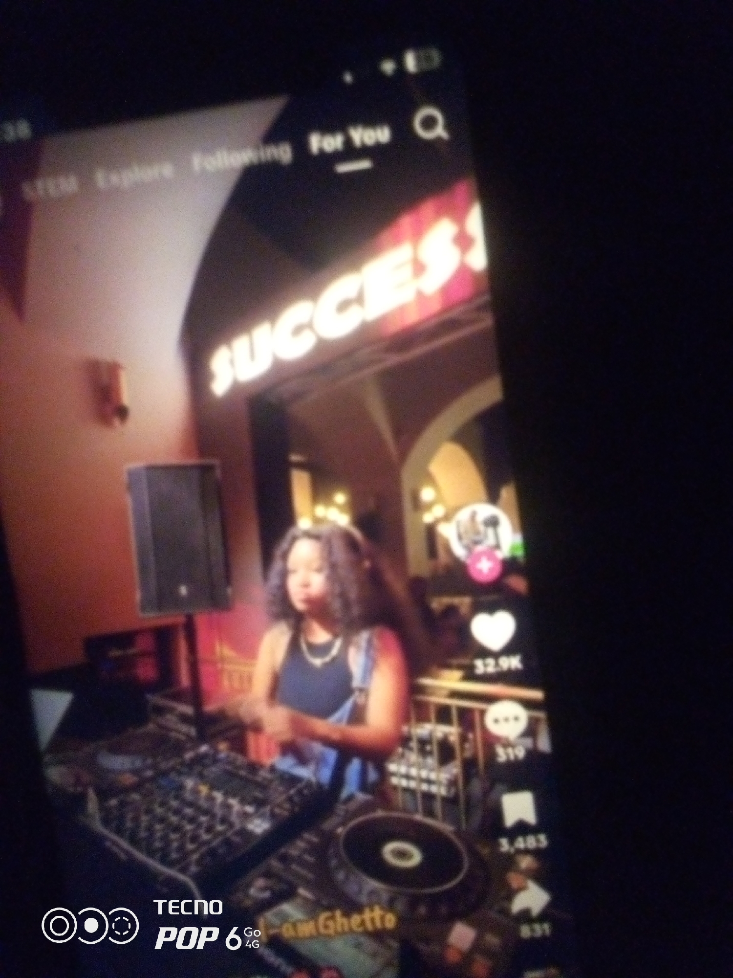 The image shows a person, likely a woman with curly hair, standing behind a DJ setup. The DJ setup consists of various electronic equipment, including turntables and a mixer. The woman is wearing a dark top and a necklace. Behind her, a large sign with the word "SUCCESS" in illuminated letters is visible. To the left of the DJ, there is a speaker on a stand. The overall environment appears to be an indoor venue, possibly a club or event space. On the right side of the image, a social media interface is visible, displaying icons for likes, comments, shares, and a profile picture with a plus sign. The phone displaying this interface is reflecting the scene. The bottom of the image shows branding for "TECNO POP 6 4G". There is no information in the image that indicates the location is Howayi, Nigeria.