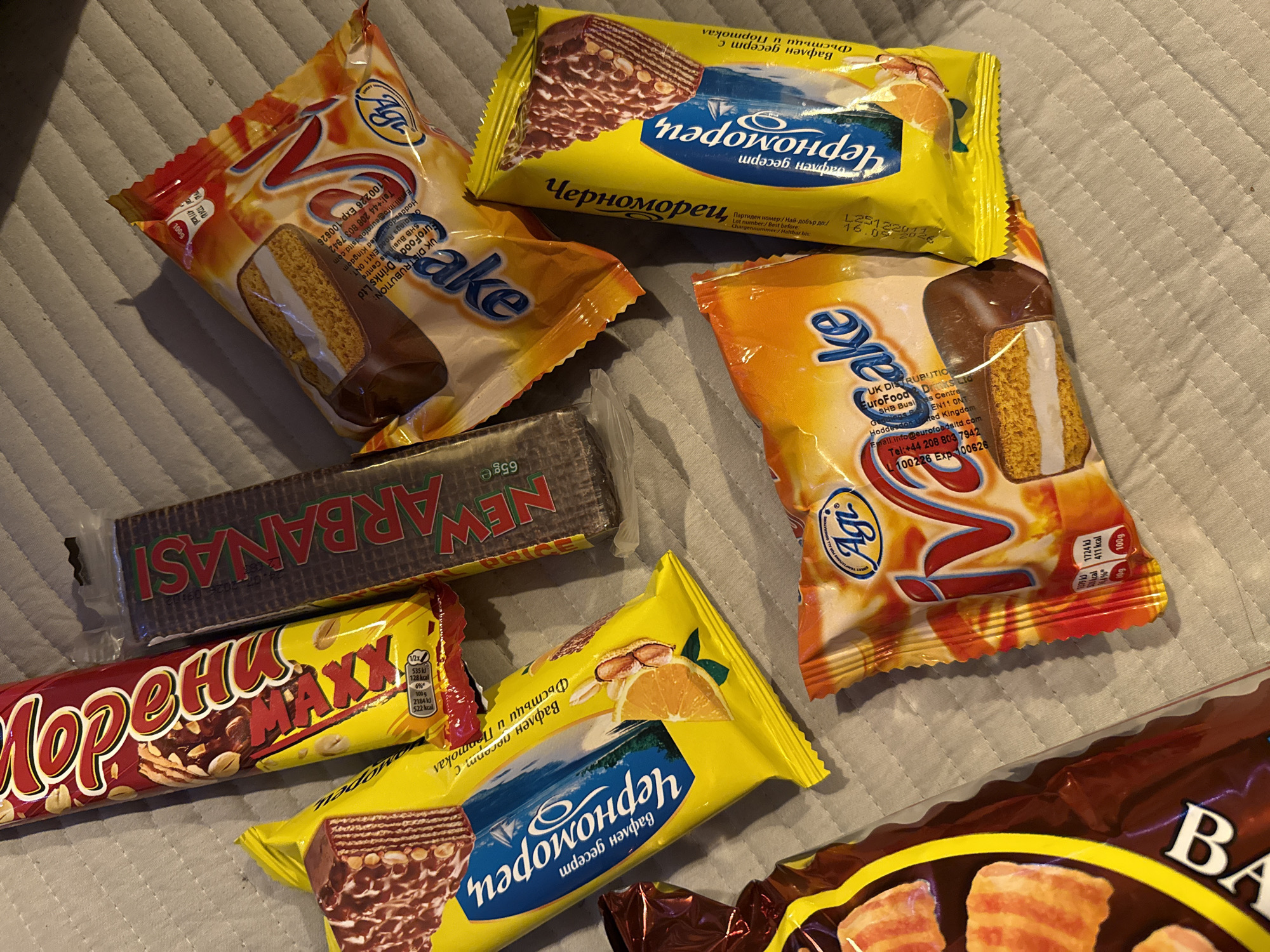 A flat lay of various confectionery items is arranged on a textured grey surface, likely a blanket or tablecloth, on a dimly lit surface. The items include a large red and yellow chocolate bar with peanuts, a dark rectangular package with "NEW ARBANASI" printed vertically in red and green lettering, and several individually wrapped yellow packages. One yellow package displays an orange slice illustration and text in Cyrillic, and appears to contain a wafer dessert with chocolate. Another yellow package, also featuring Cyrillic text and a similar wafer dessert illustration, is positioned adjacent. Two orange and yellow packaged cakes, branded "iCake," with an illustration of a cake slice and nutritional information, are also visible. A partially visible brown package with yellow lettering, featuring images of baked goods, is present in the lower left corner. The lighting casts shadows, with a prominent shadow from the upper left item falling across the arrangement. The specific location of Great Yarmouth, United Kingdom, cannot be determined from the visual information provided.