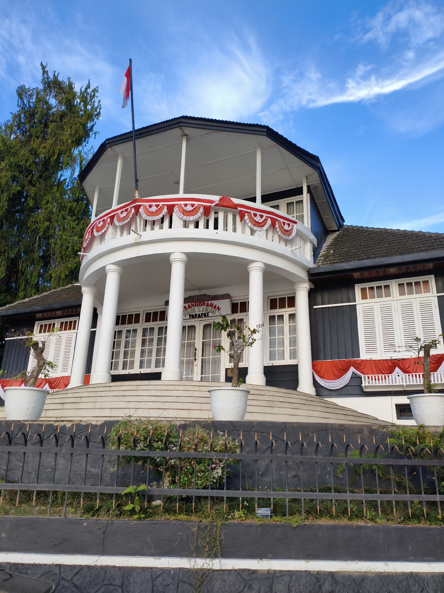 A wide-angle, eye-level shot captures the front of a colonial-style building under a bright blue sky with wispy clouds. The building is painted dark grey with white trim and shutters. A prominent white, semi-circular balcony with fluted columns supports an upper level. Red and white bunting adorns the railings of both the balcony and a lower covered porch. An Indonesian flag flies from a flagpole on the upper level. The building is set back from the viewer, with a grey metal fence in the foreground. Lush green trees are visible to the left of the building. The ground leading up to the building is covered in light grey stone steps. The text "SANGGRANHAN" and "PARAPAT" is visible above the entrance on a white sign. The image appears to be taken in the city of Tiga Raja, Indonesia, given the context.