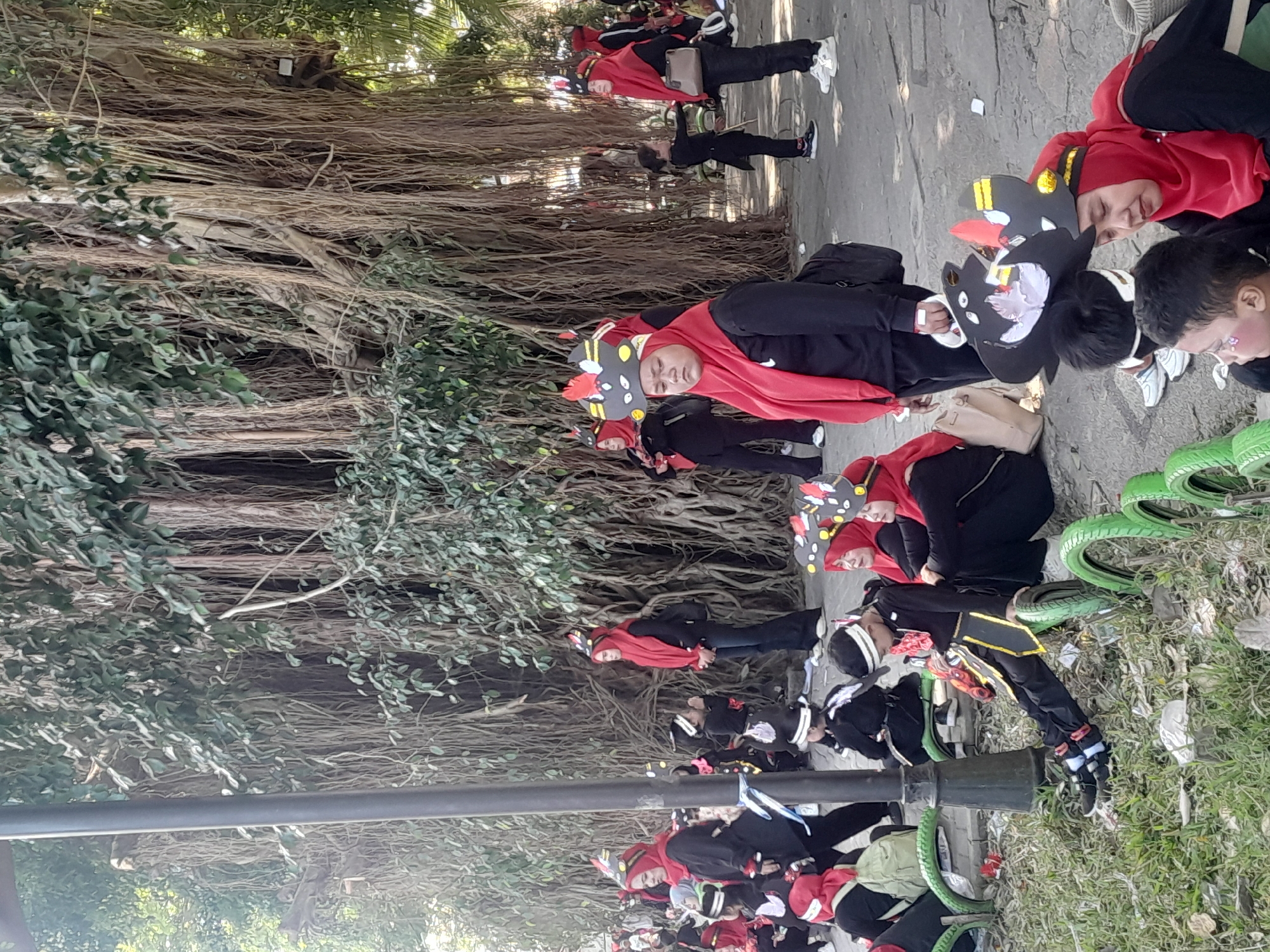 In Genteng Kulon, Indonesia, a group of people, predominantly women and children, are gathered outdoors, possibly for a parade or cultural event. They are dressed in coordinating black and red outfits, with many wearing distinctive black and yellow hats adorned with red and white accents. The backdrop features a large tree with numerous hanging aerial roots, creating a natural, somewhat dense environment. Some individuals are seated on green repurposed tire planters, while others stand or walk. A lamppost is visible on the left side of the frame. The overall atmosphere appears to be one of community gathering and cultural participation.