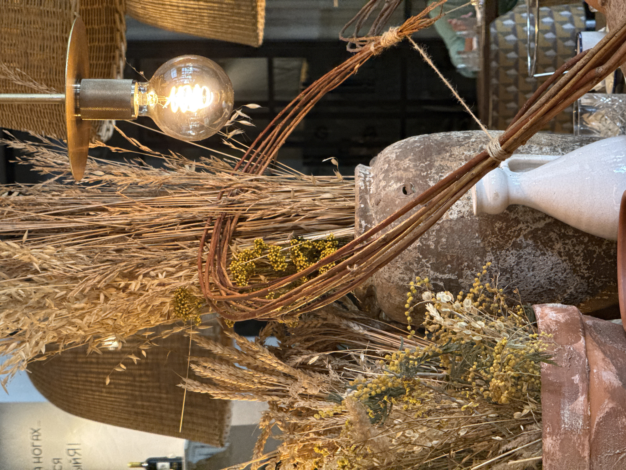 This image captures an indoor display, likely within a shop or market stall in Krasnoyarsk, Russia, featuring a rustic and natural aesthetic. The scene is dominated by a variety of dried natural elements and handcrafted objects, arranged closely together.
A vintage-style clear glass light bulb with a glowing spiral filament, mounted on a brass-colored fixture, serves as a prominent light source in the upper left. Below and around it, large bundles of dried golden-brown grasses or grains, resembling wheat or oats, create a textural backdrop. Interspersed within these are clusters of smaller, dried yellow flowers or seed pods, and some lighter, almost white, dried plant material.
Adding to the natural theme are several bundles of thin, reddish-brown twigs, tied together with natural twine, some forming a curved structure. In the background, a large, weathered, greyish-brown ceramic or clay urn with a rough texture is partially visible. Next to it rests a white ceramic jug or vase with a narrow neck, exhibiting a simple, artisanal quality. The textures of woven wicker or basketry can be seen in the background and lower left.
The warm glow of the light bulb suggests artificial lighting, creating an inviting atmosphere. The color palette is primarily earthy, consisting of various shades of brown, gold, beige, grey, and off-white, accented by muted yellow. The overall impression is one of natural warmth and artisanal craftsmanship.
Reflected on a surface in the bottom left corner, partially visible Cyrillic text can be deciphered, including the word "НОГАХ" (NOGAKH), consistent with the provided location context of Krasnoyarsk, Russia. No direct activity or people are present; the image is a still life focused on the decorative arrangement.