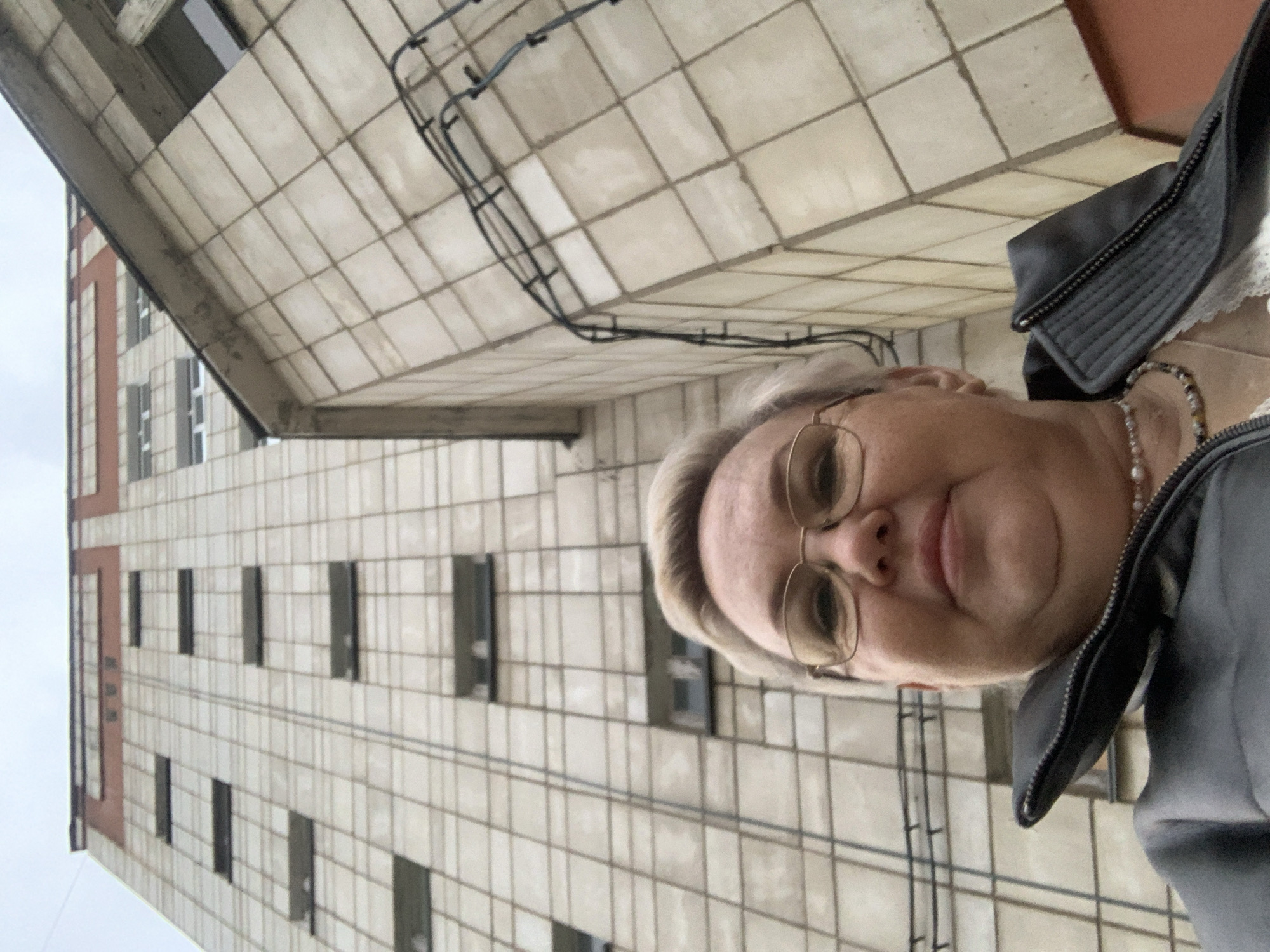 A medium shot shows a woman wearing glasses, a jacket, and a pearl necklace, positioned in front of a tall, tiled building. The building has a rectangular facade with evenly spaced windows and a vertical red stripe on the left side. Overhead, a tiled overhang extends above the woman, and wires are visible against the tiled surface. The image was taken from a low angle, suggesting the woman is looking down at the camera. The location appears to be in Chaikovsky, Russia, based on the building's architecture.