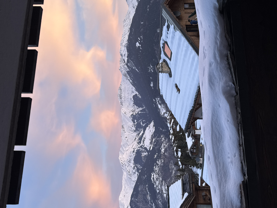 This image captures a serene winter scene in an alpine environment, likely Courchevel, France, viewed from a window or balcony. The sky, displaying soft pink, orange, and blue hues with scattered clouds, suggests either sunrise or sunset.
In the background, majestic snow-capped mountains dominate the horizon. The mid-ground features several traditional-style chalets, their roofs heavily blanketed in pristine white snow. On the roof of one prominent chalet, a small, reddish-brown animal figure, possibly a dog or fox statue, stands amidst the snow, near a rectangular, light-colored object that could be a skylight. Another chalet in the distance shows a partially visible person in a blue jacket on a balcony or upper floor.
The foreground includes dark structural elements on both sides: a window or balcony frame on the left, and a thick layer of snow on a ledge or railing on the right, with a dark wooden frame adjacent to it. Faint footprints or animal tracks are visible in the snow on this foreground ledge. The overall activity is a static, picturesque view of a snowy landscape. There is no visible text in the image.
