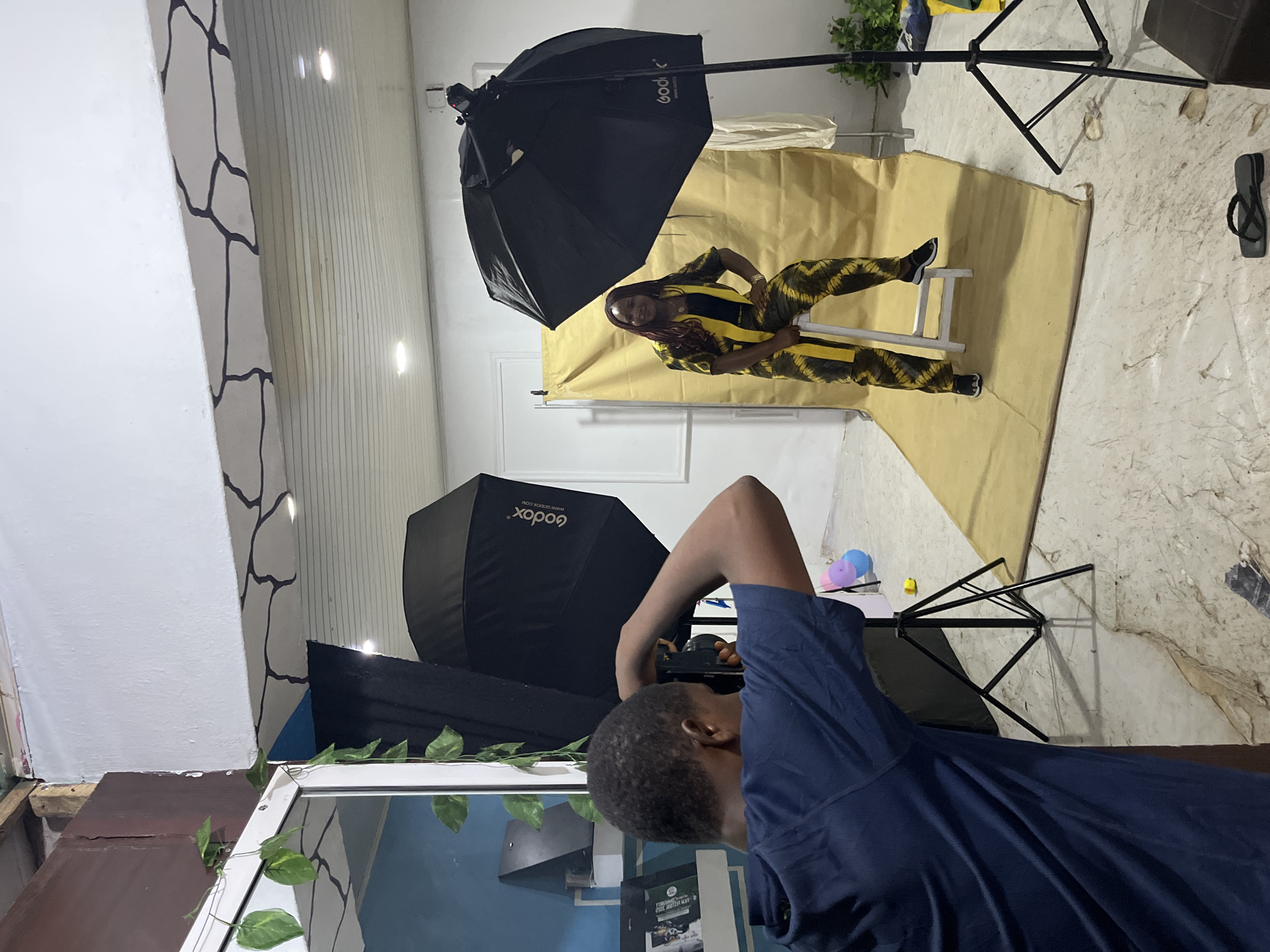 An indoor photography studio in Jos, Nigeria, is depicted with two individuals engaged in a photoshoot. In the foreground, positioned to the left and seen from the back, a male photographer with short dark hair, wearing a dark blue short-sleeved top, holds a black DSLR camera to his eye, actively photographing. To the right, in the mid-ground, a female model with long, dark braided hair (featuring reddish-brown highlights) sits on a simple white backless stool, posing. She is dressed in a yellow and black patterned two-piece outfit, likely tie-dye or batik, and wears black sneakers with white soles. Her left hand rests on her hip, while her right arm is relaxed on her thigh; she looks directly towards the perspective of the original photographer.
The studio setup includes two black Godox photography strobe lights with softboxes on stands, illuminating the model. One large square/octagonal Godox softbox is positioned to the left of the model, and another umbrella-style Godox softbox is to her right. The backdrop is a large, slightly wrinkled sheet of yellow-beige fabric, extending from the wall onto the floor.
The environment features light-colored, possibly tiled, flooring with irregular patterns. The walls are predominantly white, with a section above the model displaying a decorative black crack-like pattern. Exposed wooden beams are visible in the top left corner, suggesting an unfinished or repurposed space. A blue wall is visible through a doorframe or reflection on the far left. Recessed spotlights are visible in the white ceiling. Artificial green vine plants are draped around a white doorframe on the left. On a shelf behind the photographer, dark objects, possibly books or props, and a black triangular stand are visible. A single black flip-flop lies on the floor near the model's feet. Two partially deflated balloons, one pink and one blue, along with a small yellow object, are near the base of the left light stand.