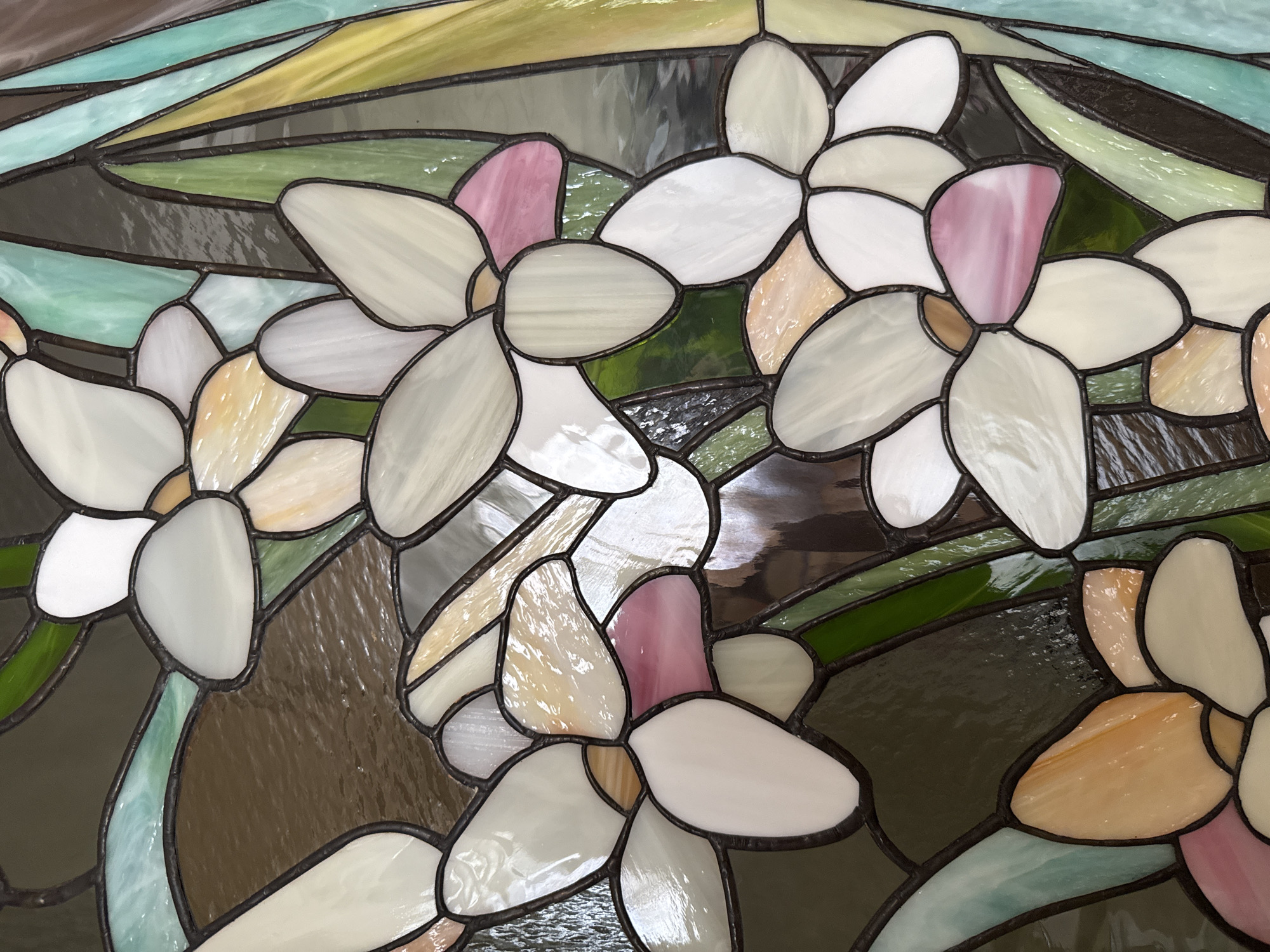 The image shows a close-up of a stained-glass panel with a floral design. The flowers have white and pastel-colored petals, outlined with dark lead lines. The background consists of geometric shapes filled with various shades of green, yellow, brown, and grey glass. The overall style is reminiscent of Art Nouveau. The location is unknown, and there is no evidence to suggest this photo was taken in the city of Vsevolozhsk, Russia.