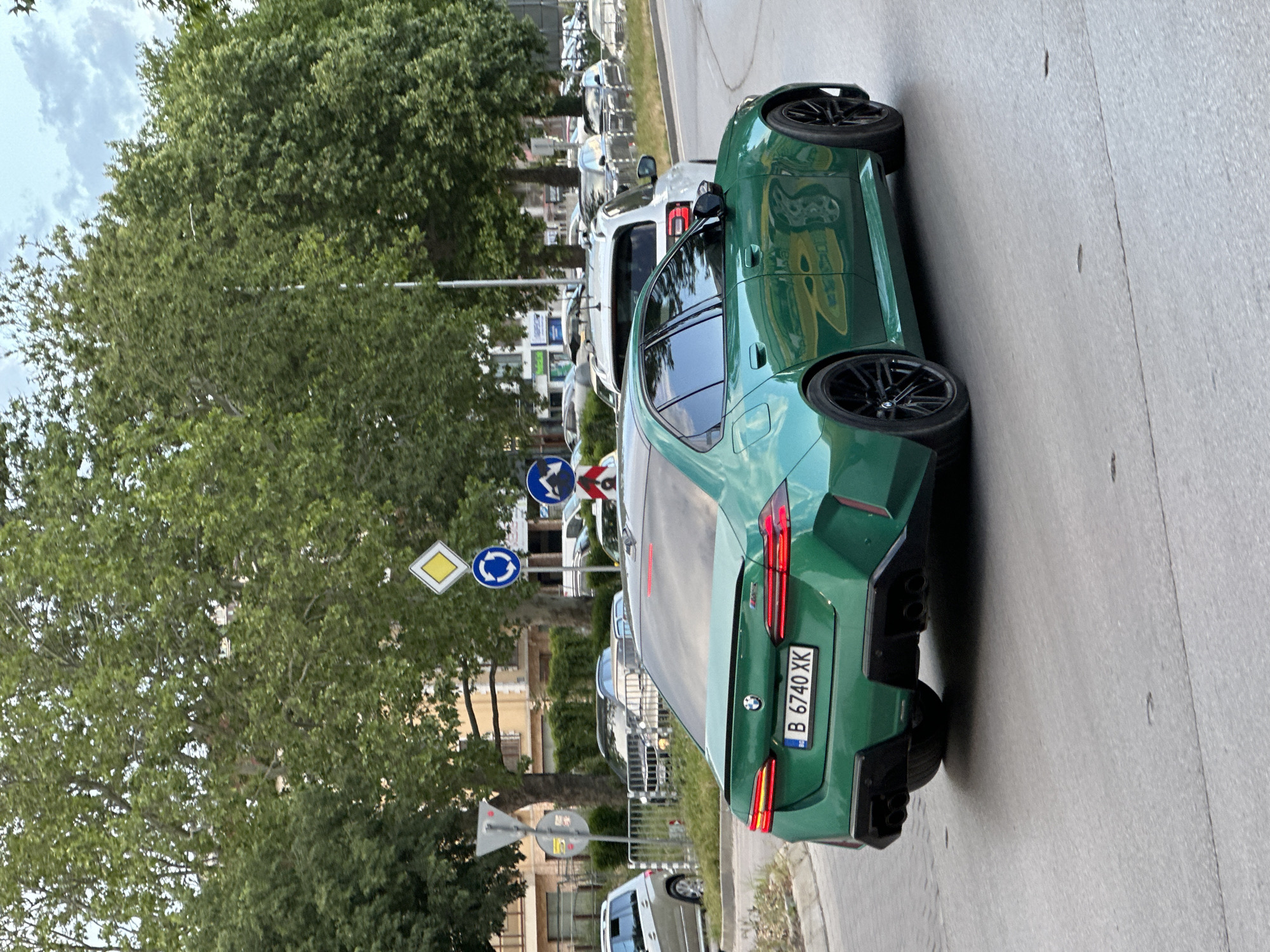 A green BMW M3 with Bulgarian license plate B 6740 XK is parked on a street in Varna, Bulgaria. It is parked near a roundabout and a "give way" sign. The street is lined with trees, and there are other cars parked nearby. The sky is cloudy.