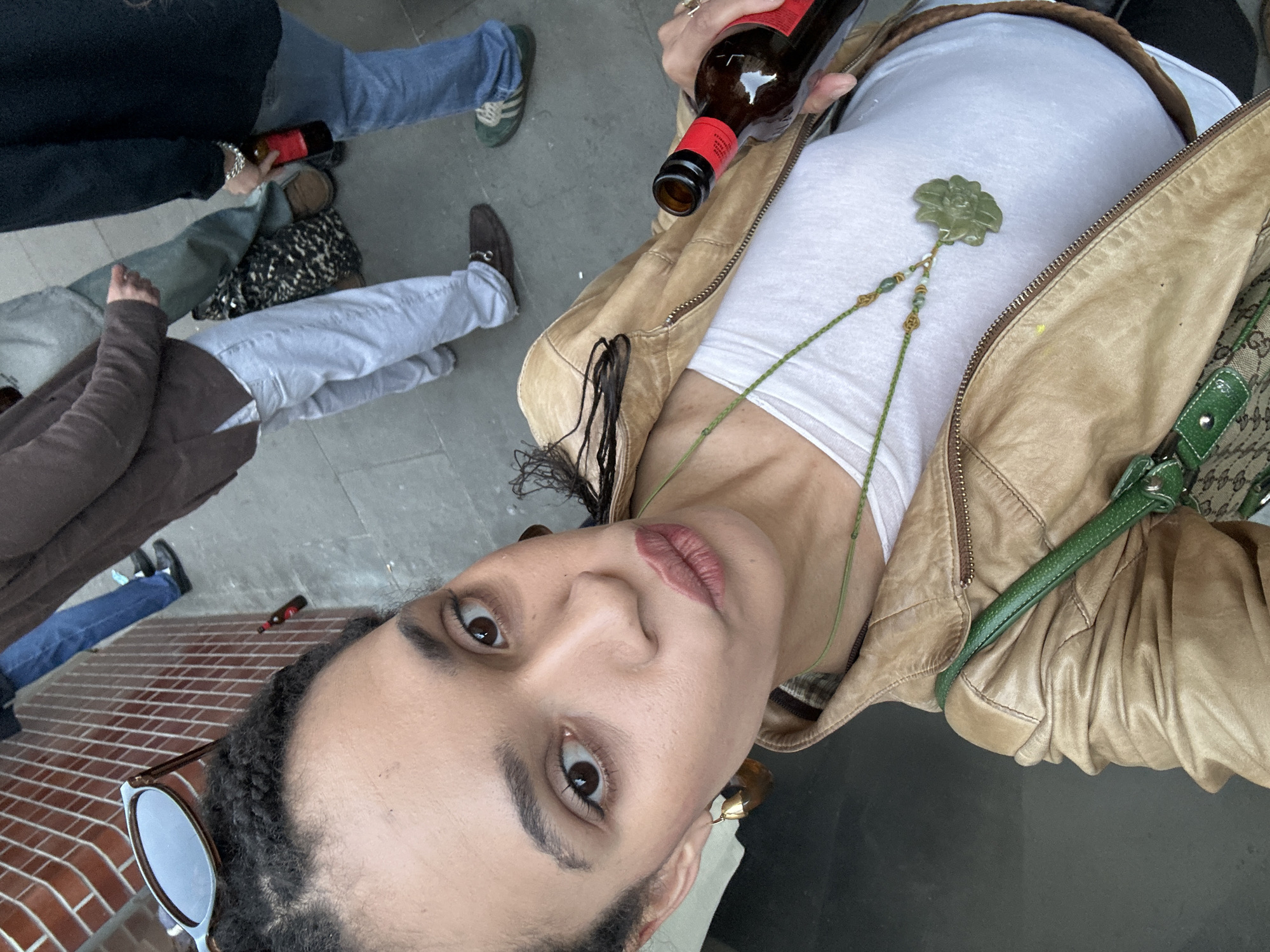 A tilted, low-angle selfie captures a person with dark braided hair, light brown skin, and dark eyes looking directly at the camera with a neutral expression. They are wearing a white t-shirt, a tan-colored leather-look jacket with a zipper, and a green beaded necklace adorned with a light green floral pendant. Light brown lipstick is visible on their lips. In their right hand, they hold a dark glass bottle with a red label. A pair of sunglasses rests on their head or hair, and a brown earring is visible on their right ear. A shoulder bag with a green strap and a patterned fabric is partially seen to their right.
The setting appears to be an outdoor urban environment, consistent with a street or public space in London, United Kingdom, during daylight hours. The background features the lower halves of several other individuals, suggesting a casual gathering. One person wears dark pants and holds a similar red-labeled bottle. Other visible legs wear light blue jeans paired with brown loafers, and light blue jeans with green and white striped sneakers (possibly Adidas). A leopard-print bag is visible on the ground near a person's feet. The ground surfaces consist of grey concrete and some brick paving. The overall mood is relaxed and informal. No specific brands or text are clearly legible on any items in the image.