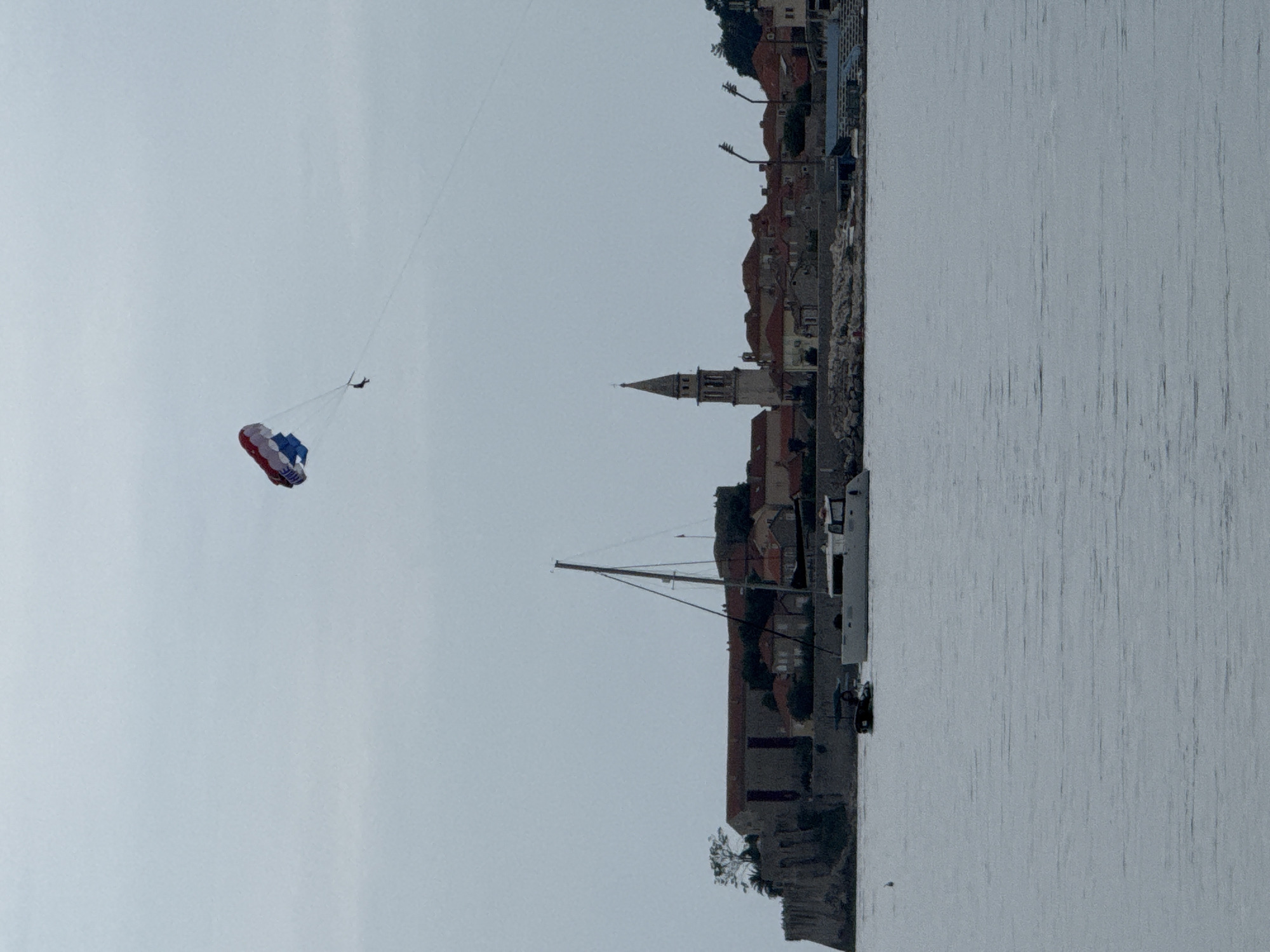 The image shows a cityscape with a body of water in the foreground, potentially part of Budva, Montenegro. The city includes buildings with red roofs and a church spire. A sailboat is visible near the shore. In the sky, a parasail is being towed with a person suspended. The overall scene suggests a recreational activity on the water near an urban area.