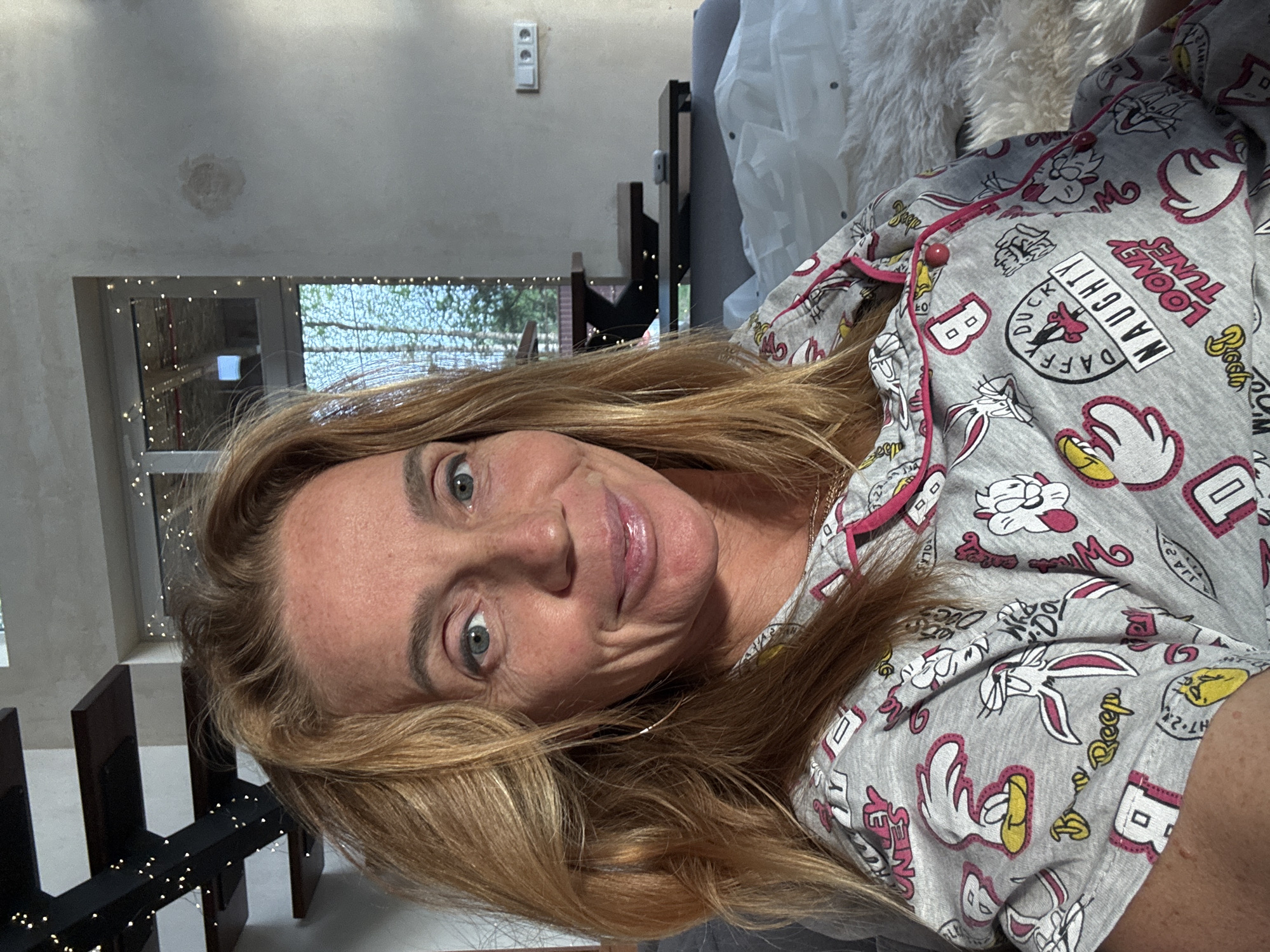 A woman is seen indoors, likely in a residential setting. She has light blonde hair and is wearing a gray pajama top patterned with cartoon characters, including the inscription "Naughty Looney Tunes." In the background, there's a window adorned with lights, and a dark railing is visible near her. A light-colored wall and some furniture, including what appears to be a gray sofa with a fluffy white throw, are also present in the background. Based on the available visual evidence, there's no definitive information to confirm that the location is in the city of Steklyannyi, Russia.