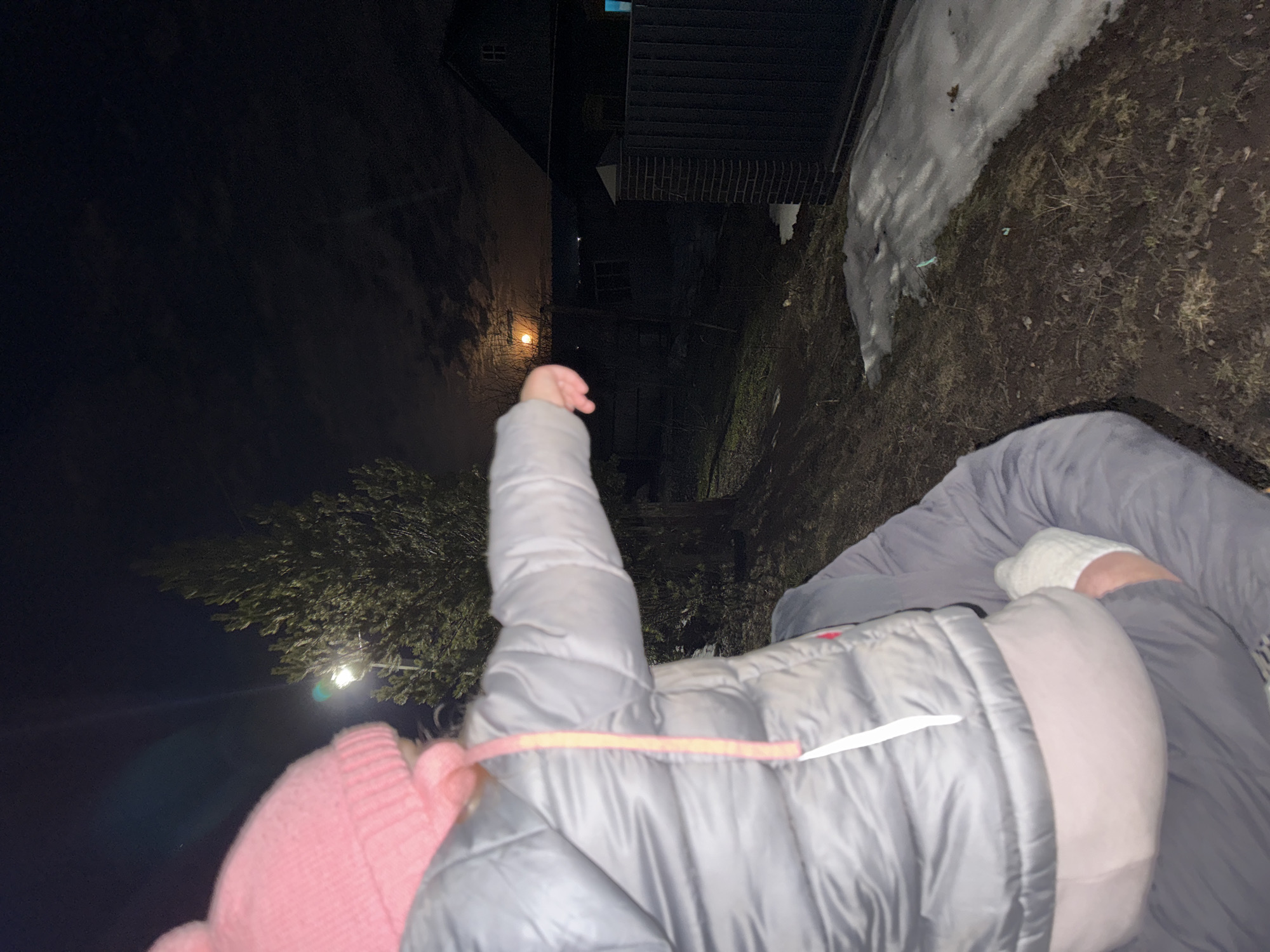 A person lies on the ground covered in a gray comforter, wearing a pink knitted hat. The photo appears to be taken at night in an urban setting, with buildings and trees visible in the background. The specific location is Горный, Russia, with snow patches indicating a cold climate.