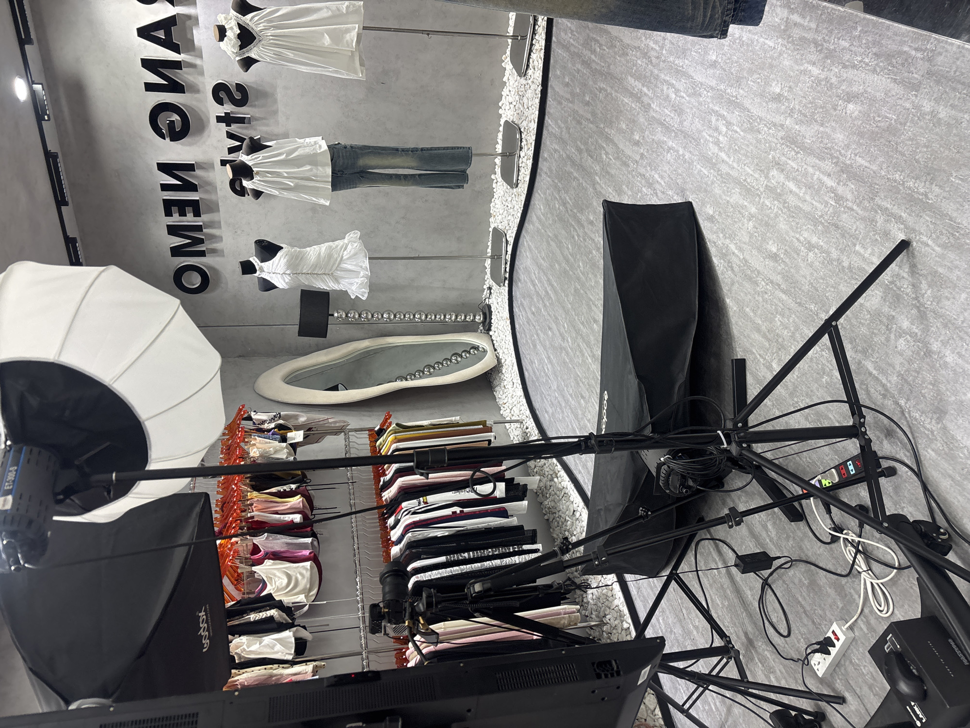 An indoor studio setup displays clothing on mannequins and racks, with photographic equipment in the foreground. Three mannequins are dressed in white tops, one with a ruffled dress, and another in flared jeans. Racks of various colored garments are visible behind them. A large softbox light and camera equipment, including a tripod and monitor, are positioned in front of the displays. The floor is a textured grey surface, with small white pebbles decorating the area around the mannequins. A large, ornate mirror stands to the left of the mannequins. The wall behind the mannequins features large, black, three-dimensional letters that spell out "OMEN ONIA". The overall scene suggests a professional setting for product photography or a fashion retail display.