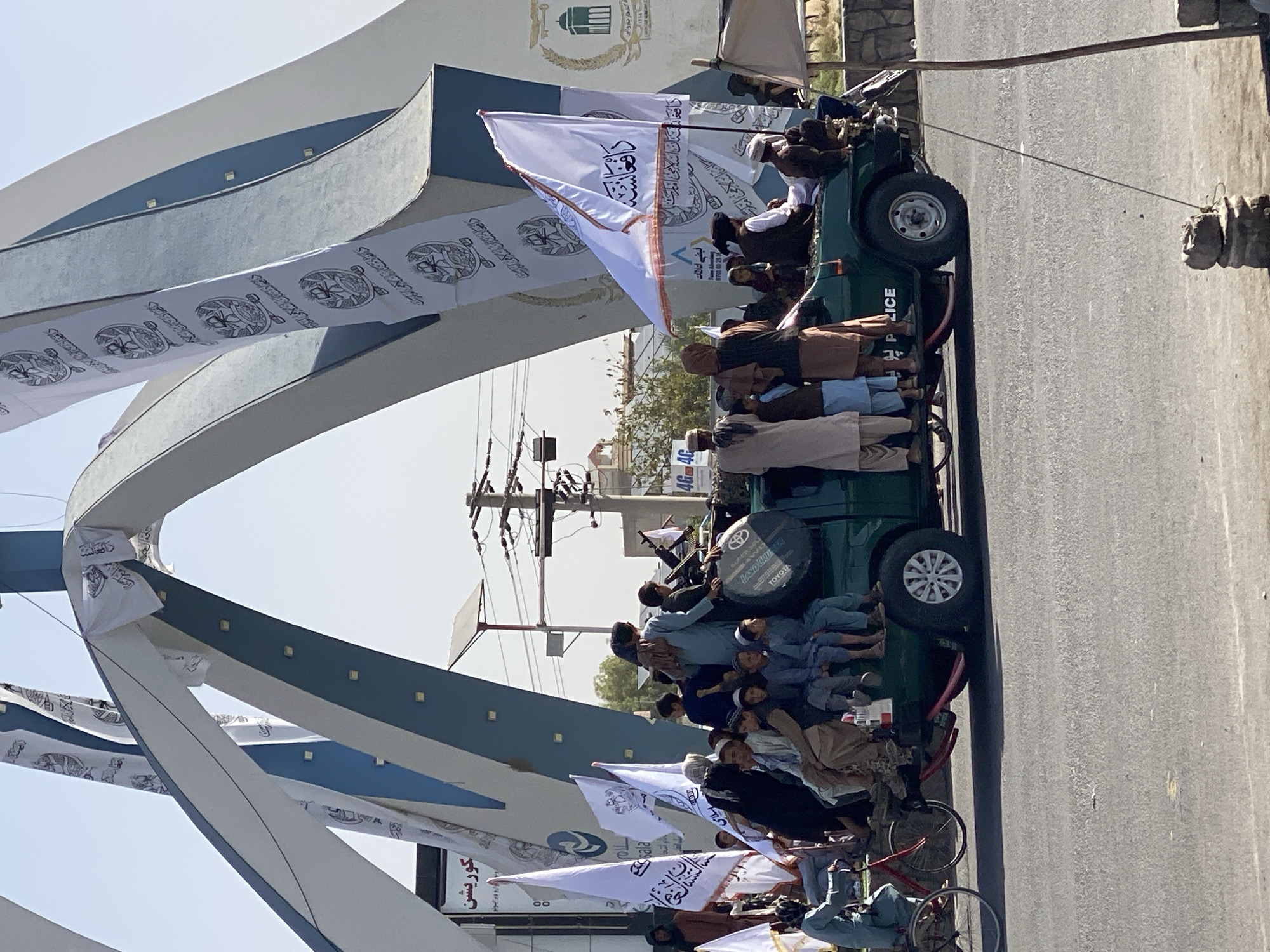 The image depicts a scene in Kandahar, Afghanistan, featuring a parade or procession. In the foreground, a dark green pickup truck is loaded with numerous people, primarily young men and boys, some of whom are seated on the truck bed and others standing. They are dressed in traditional Afghan attire, including shalwar kameez and turbans. Several white flags with Arabic script and emblems are prominently displayed, attached to poles and held by individuals. A large, arched monument with blue and white accents, adorned with similar white banners, is visible in the background. The environment appears to be urban, with paved roads, streetlights, and buildings. The overall atmosphere suggests a public gathering or celebration.