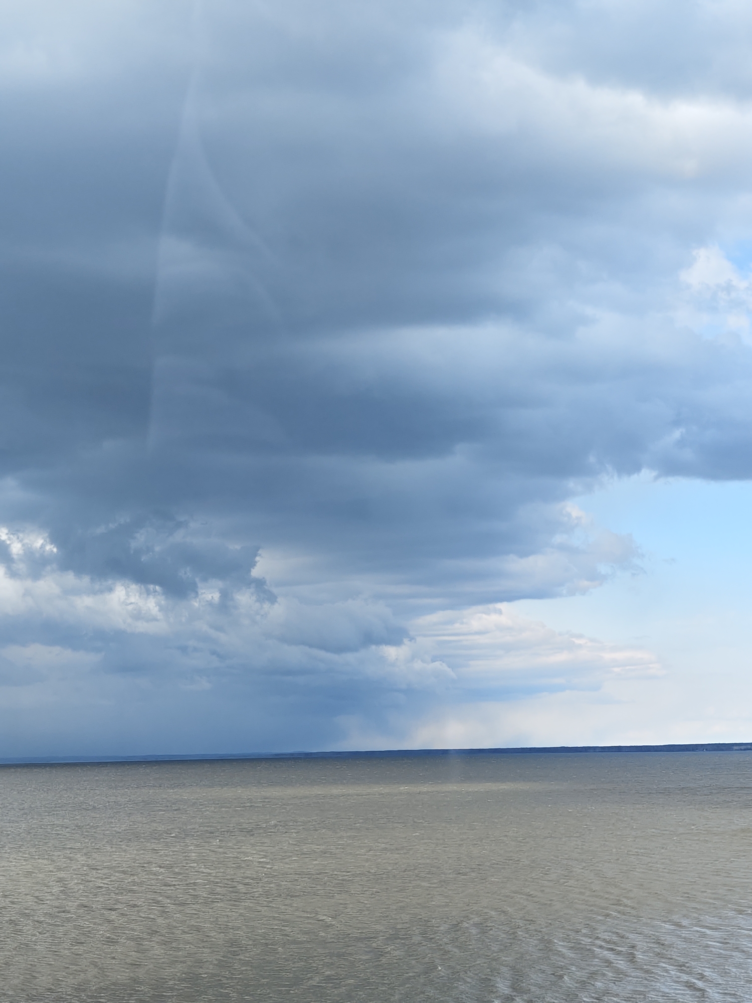 The image presents a vast natural landscape, dominated by a large body of water under a dramatic, heavily clouded sky. The upper half of the frame is filled with thick, dark grey storm clouds, appearing heavy and turbulent. A small section of lighter blue sky is visible in the upper right quadrant, offering a contrast to the prevailing overcast conditions.
A notable weather event is unfolding in the distance over the water, where visible shafts of rain descend from the dark clouds, particularly towards the left, indicating active precipitation. Adding to the atmospheric conditions, a very faint and translucent arc, possibly a partial rainbow or another atmospheric optical phenomenon, is discernible within the clouds in the upper left section, arching downwards from the top edge.
The lower half of the image features a wide expanse of water, likely a lake or a large river, consistent with the Chaikovsky, Russia location and its proximity to the Votkinsk Reservoir. The water surface is a muted grey-green, showing subtle ripples and reflecting the somber sky. A distant, dark horizon line, possibly indicating land, stretches across the width of the image where the water meets the sky. The time of day appears to be daytime, under stormy or pre-stormy conditions. No people, man-made objects, or visible text are present in the image. The scene evokes a sense of expansive, brooding natural beauty.
