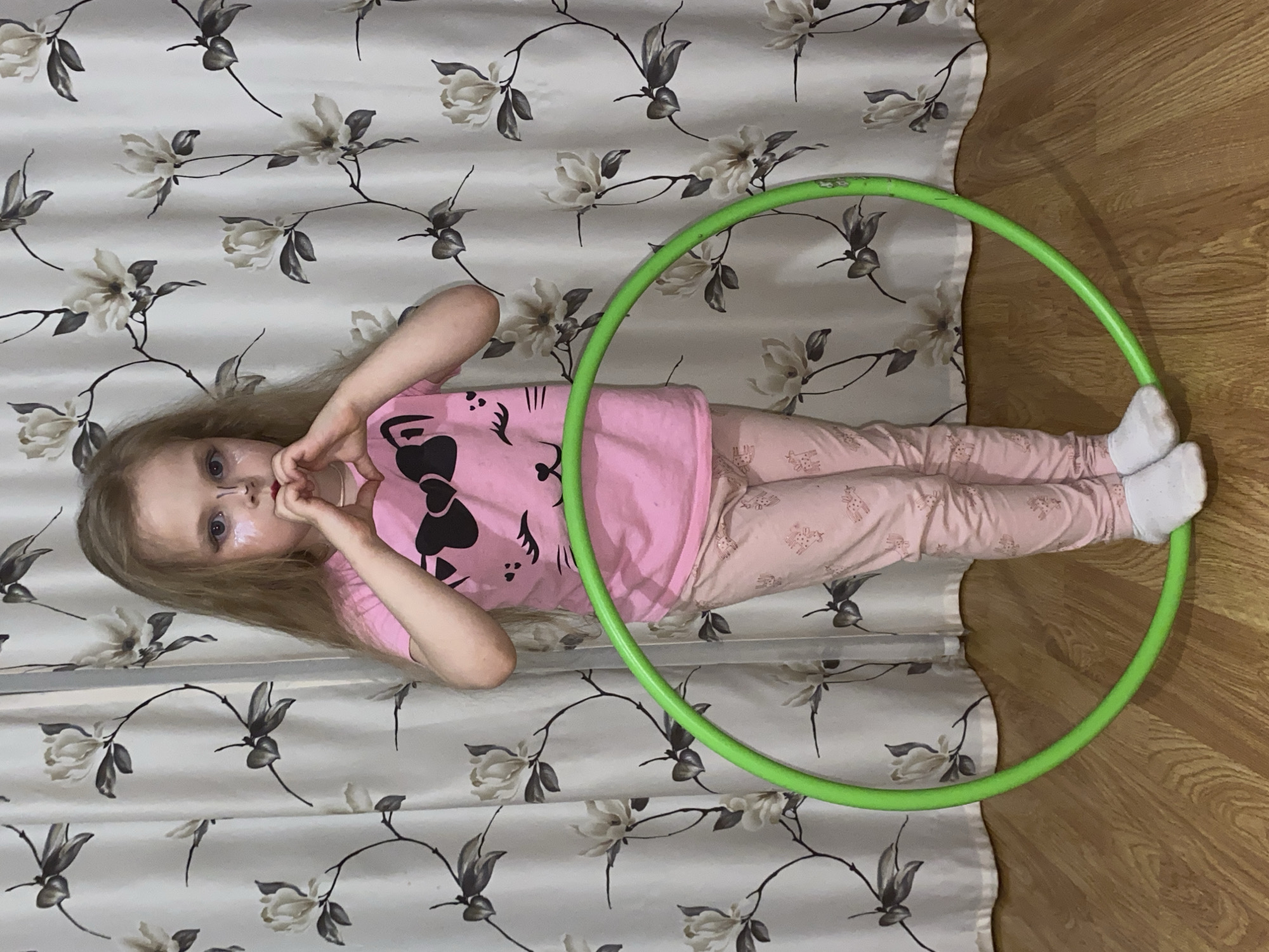 A young girl is depicted in an indoor setting, posing for the camera while standing inside a bright green hula hoop. She has long, straight blonde hair and blue eyes, and there appears to be light-colored makeup or face paint around her nose and upper lip. She is making a heart shape with her hands near her face, looking directly at the viewer with a neutral to slightly playful expression.
The girl is wearing a pink short-sleeved t-shirt with a black design on the front, resembling a cat face with a large bow, possibly from the Hello Kitty brand. Her pants are also pink, featuring a subtle pattern of light-colored motifs, possibly small animals like giraffes. She is wearing white socks.
The background consists of a light-colored curtain or fabric wall covering with a distinctive floral pattern. This pattern includes light beige or off-white flowers with dark gray or black stems and leaves, arranged in a repeating vertical design. The curtain has visible vertical folds. Below the curtain and around the girl's feet, a light wooden floor with visible grain is present. The lighting suggests an indoor environment.
This scene captures a moment of childhood playfulness. The provided context indicates the photo was taken in Votkinsk, Russia. There is no visible text in the image.