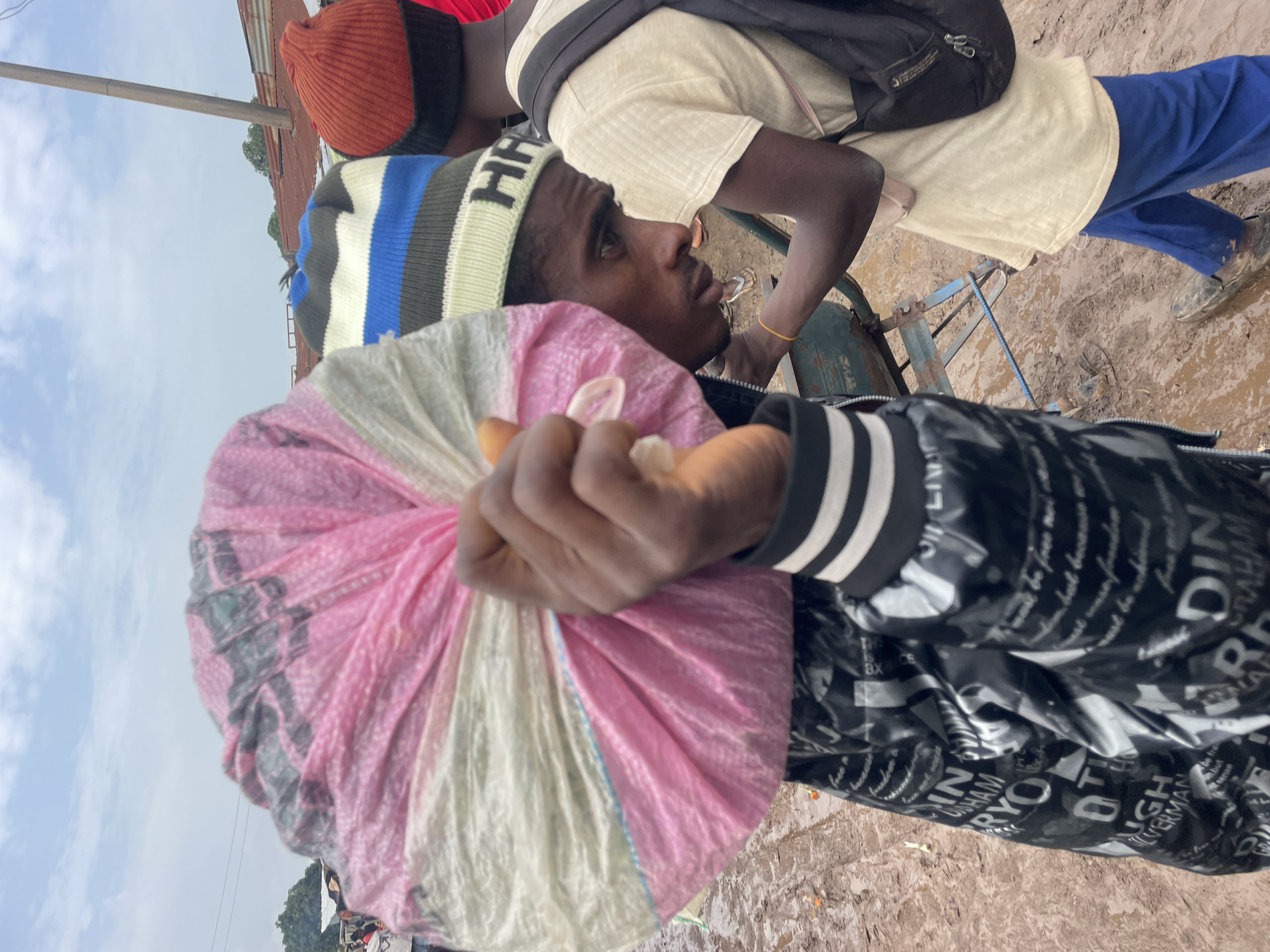 In Gura Top, Nigeria, a young man with a dark complexion and a light brown beard carries a large bundle of pink and white plastic bags over his right shoulder. He wears a black jacket with white stripes on the cuffs and a blue, white, and black striped beanie. He is walking on a muddy, uneven dirt path. To his right, another young man, also with dark skin, wears a cream-colored t-shirt, blue trousers, and an orange beanie. He has a black backpack slung over his left shoulder and is looking down at a cart. The cart appears to be made of metal with blue accents and has two large wheels. The ground is a mix of mud and debris. In the background, several more people are visible, along with buildings with corrugated metal roofs, suggesting a market or informal settlement. The sky is overcast with scattered clouds.