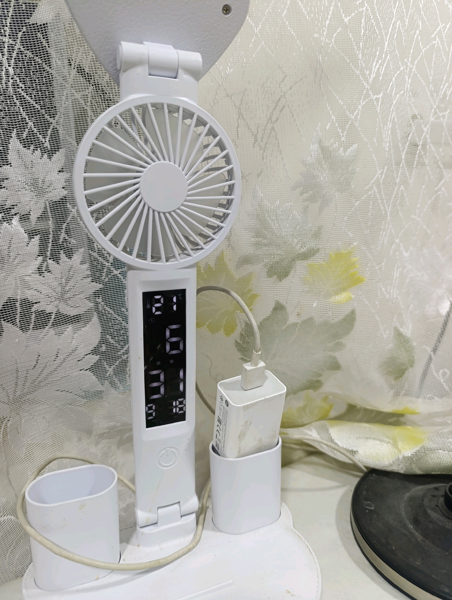 A white, multi-functional desk fan is positioned on a white surface, likely a desk. The fan has a circular head with blades and a digital display on its vertical body. The display shows "21°C" at the top, followed by what appears to be time or a date, displaying "06" and "31" and then "9:18". A white USB charging brick is plugged into the fan and rests in a white holder to the right of the fan. To the left of the fan, another white holder is visible, with a beige cable coiled around its base. The background consists of a sheer white curtain with a subtle floral pattern. The scene is indoors, with the lighting suggesting it's daytime. The presence of the fan, charging brick, and curtain indicate a domestic or office setting. No people are visible in the image. The location is Navoiy City, Uzbekistan.