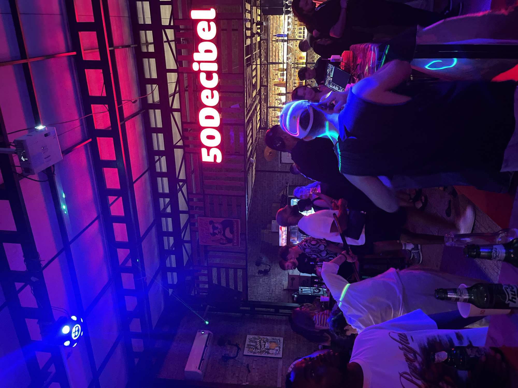 The image shows the interior of the "50Decibel" bar in Đà Nẵng, Vietnam. The bar is dimly lit with neon lights. A crowd of people stands at the bar counter, with bottles and drinks visible. Some individuals are interacting and conversing. The bar's structure consists of exposed beams and brick walls with mounted posters. A projector and a rotating blue light are affixed to the ceiling. The bar is labeled with a large, illuminated "50Decibel" sign.