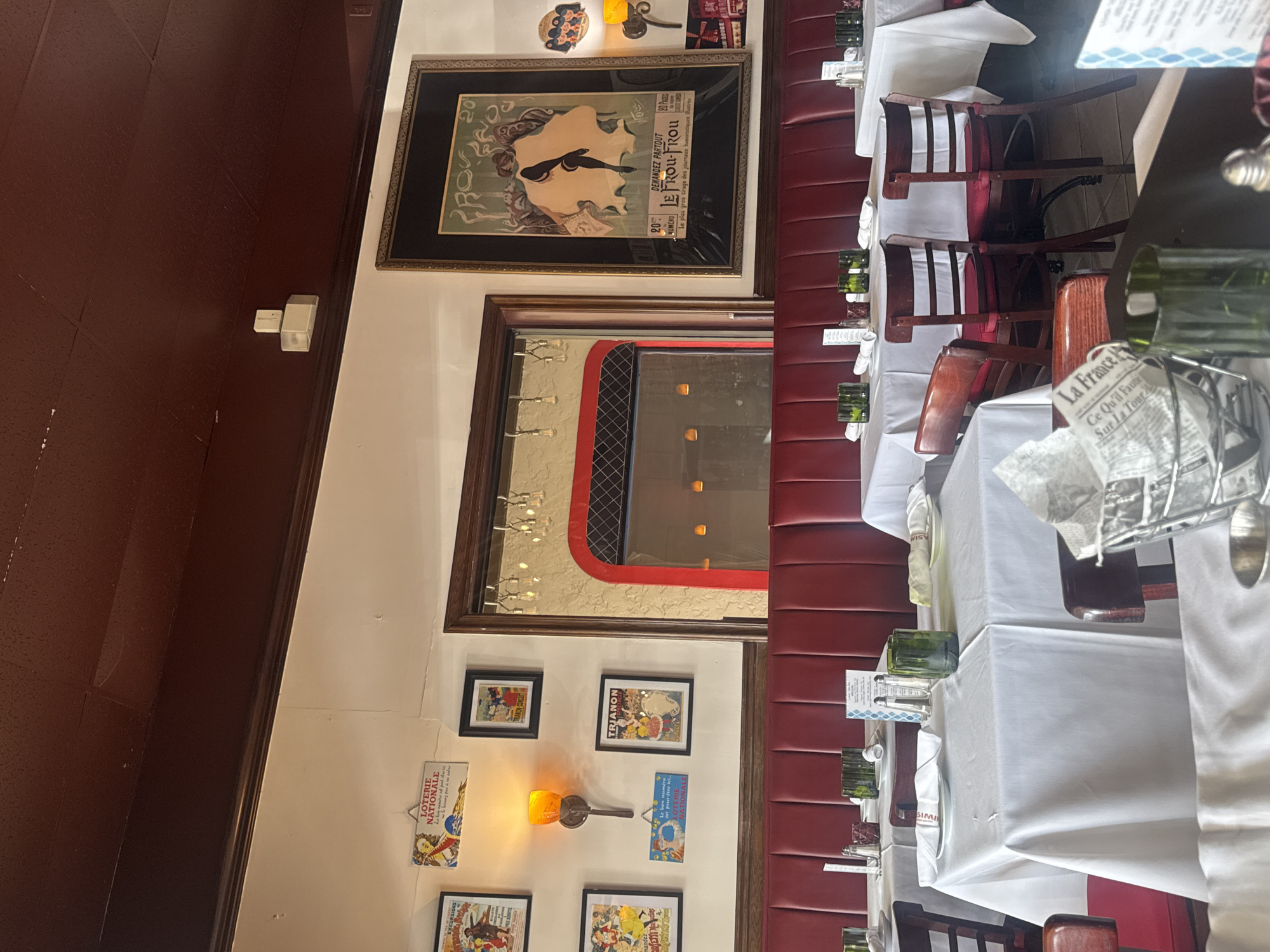 The image depicts the interior of La France, a restaurant located in Boca Raton, Florida, United States of America. It showcases tables with white tablecloths set for dining. Chairs with red seats and wooden frames are arranged around the tables. A red cushioned banquette lines the wall. Decorative elements include framed pictures and advertisements hanging on the white wall. A large framed poster with the words "LE FROU-FROU" and a woman's profile is featured above a framed mirror that reflects the interior of the restaurant. The walls are predominantly white, with the upper section painted in a dark brown color.