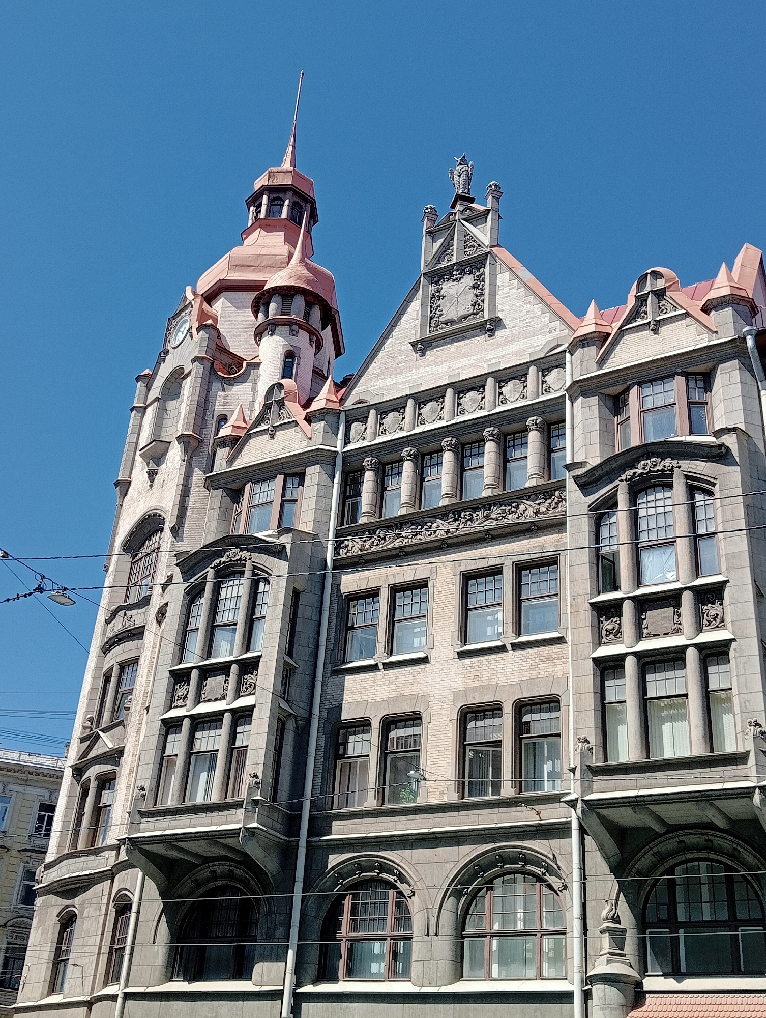 The image depicts the eclectic architecture of the Singer House (Dom Knigi), a prominent landmark in Saint Petersburg, Russia, located on Nevsky Prospekt. The building features a distinctive corner tower with a copper dome and spire, as well as a detailed facade with numerous windows, balconies, and ornamental sculptures. The architecture is a blend of Art Nouveau and historicist styles, characterized by its elaborate stonework and the use of light-colored brick. The sky above is clear and blue, indicating a sunny day. Overhead power lines and streetlights are visible in the foreground, suggesting an urban setting. No people are visible in the frame.