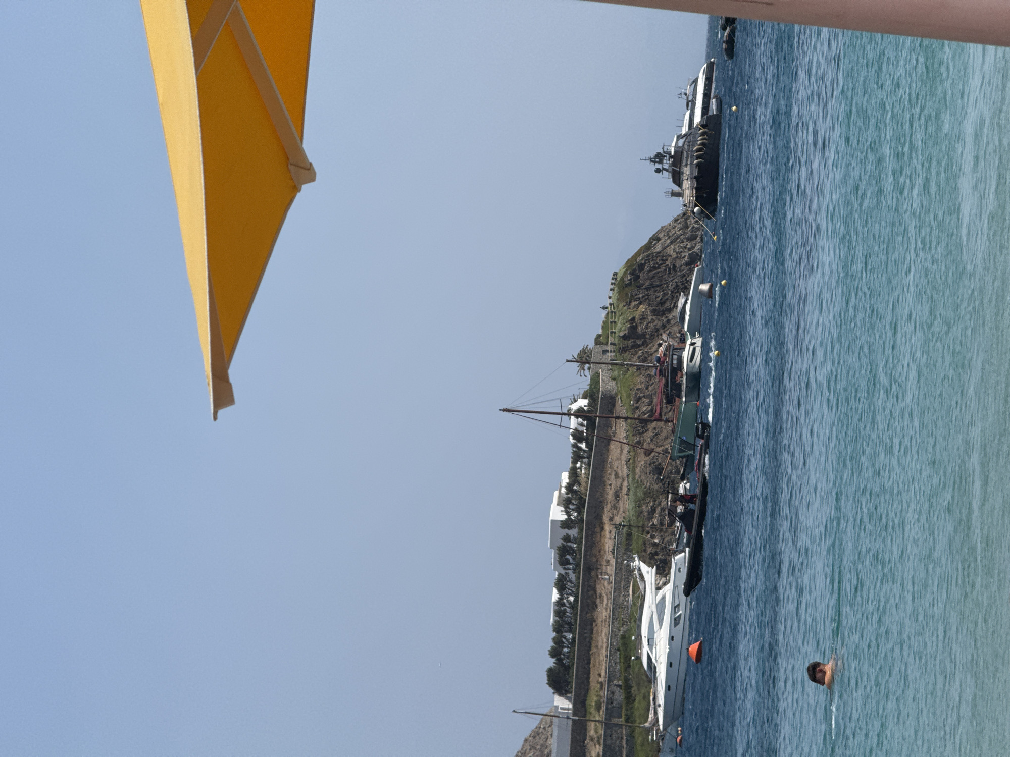 A bright yellow umbrella is partially visible in the foreground of an ocean scene. The water is a clear turquoise, with small waves. A person is swimming in the water, partially submerged. In the background, several boats are anchored offshore, including a large white yacht, a smaller black inflatable boat, and a dark gray motor yacht. Behind the boats, a rocky hillside is lined with trees and white buildings. The sky is a clear, pale blue.