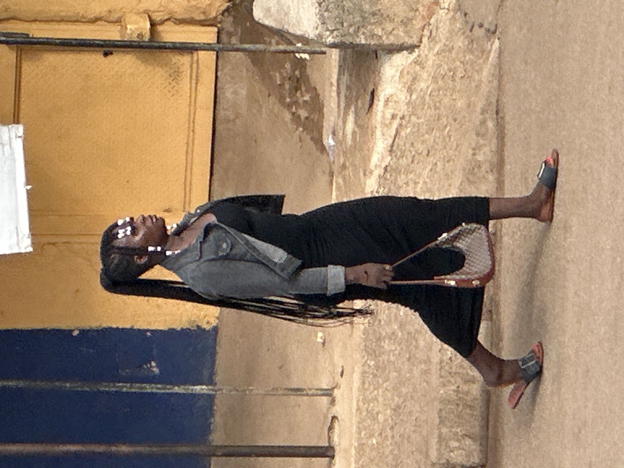 A person walks with a high ponytail and a black dress in a Nigerian city. The person is wearing a gray jacket over the dress, and carrying a purse. The person is also wearing sandals. The background of the image shows a blue wall and a yellow wall, and a fence. The ground is made of concrete.