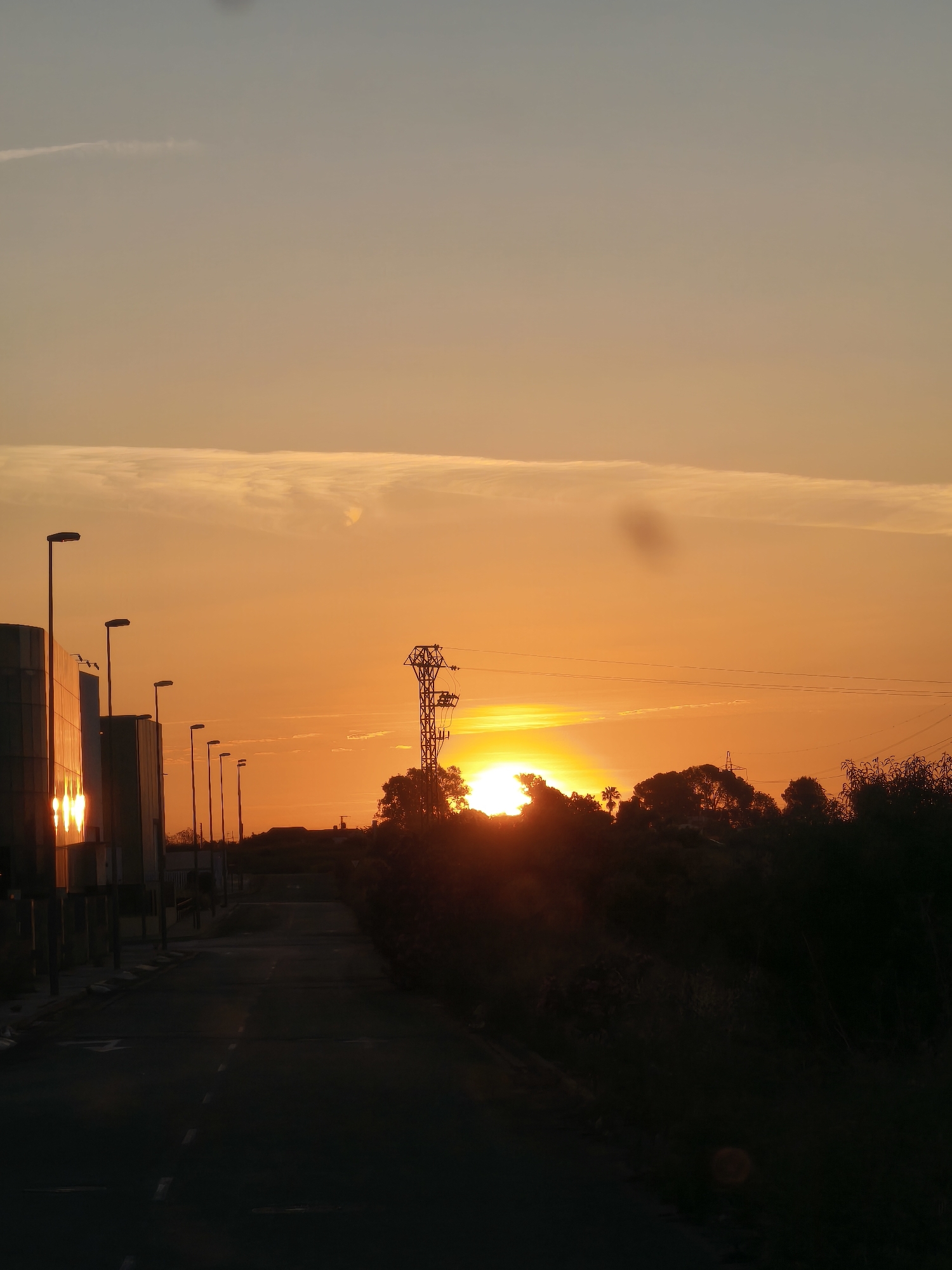 The image depicts a sunset scene on a road in Nàquera, Spain. A straight road stretches into the distance, flanked on the left by modern buildings with vertical metal slats and streetlights. On the right, trees and bushes form a silhouette against the setting sun. An electrical tower stands in the center, partially obscuring the sun. The sky is a gradient of orange hues, brighter near the horizon and fading into a pale blue towards the top. Buildings on the left side reflect the sunset glow.