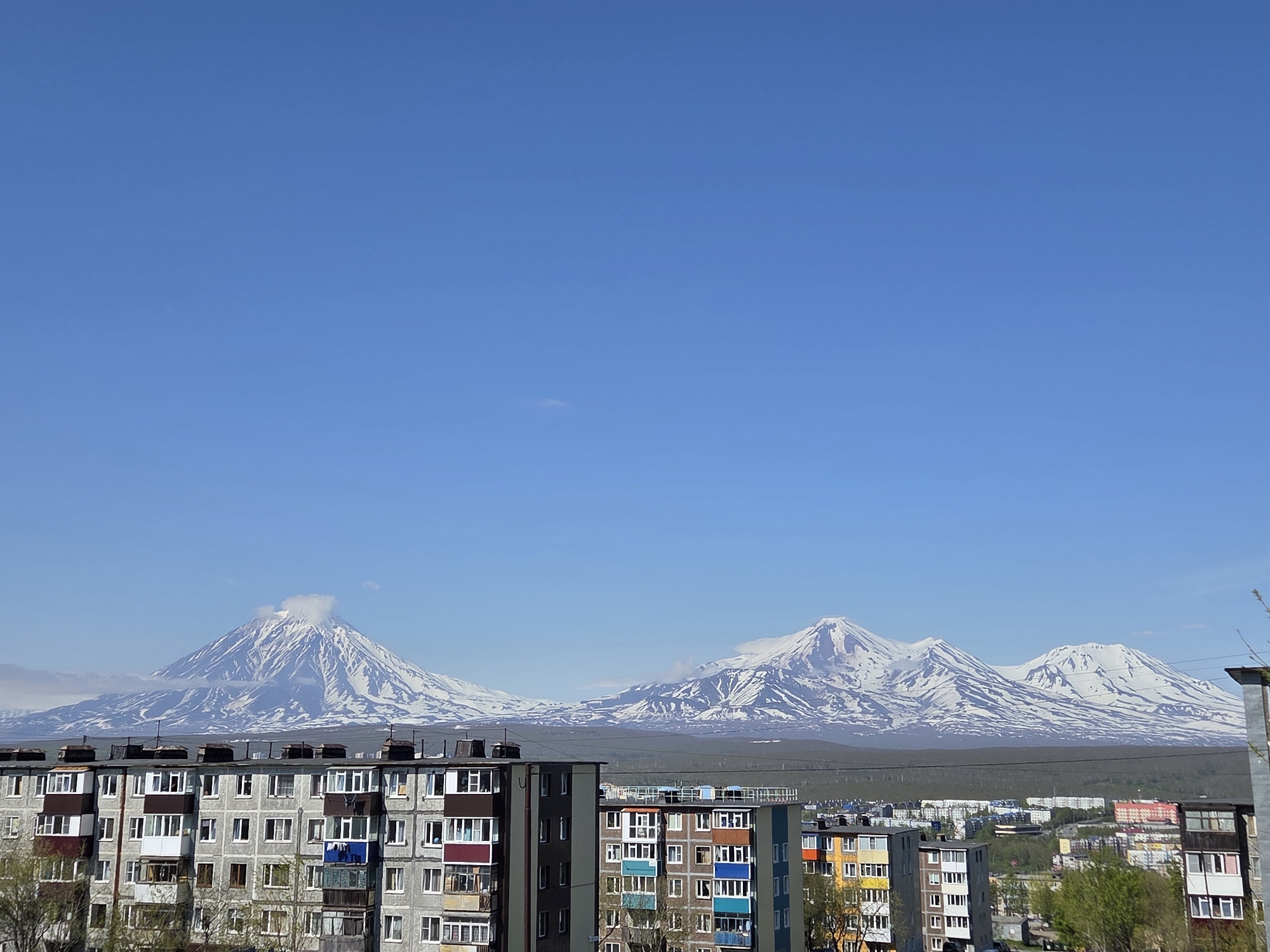 This is a landscape photograph taken from a high vantage point overlooking a city and a mountain range in the distance. The sky is a clear, bright blue. In the foreground, the tops of several multi-story apartment buildings are visible. These buildings appear to be made of concrete and have many windows and balconies. Some of the buildings are gray, while others are painted in various bright colors such as yellow, blue, and orange. The windows are rectangular, and some balconies are enclosed. Laundry can be seen hanging on a balcony in the center of the frame.
In the middle ground, there is a wide expanse of trees and smaller buildings, suggesting a sprawling urban or suburban area. Beyond this, a dramatic mountain range dominates the horizon. These mountains are snow-capped and appear to be volcanic in origin, with some showing plumes of smoke or clouds emanating from their peaks. The snow cover is extensive, highlighting the rugged contours of the terrain. The overall impression is a juxtaposition of urban development against a grand natural landscape.