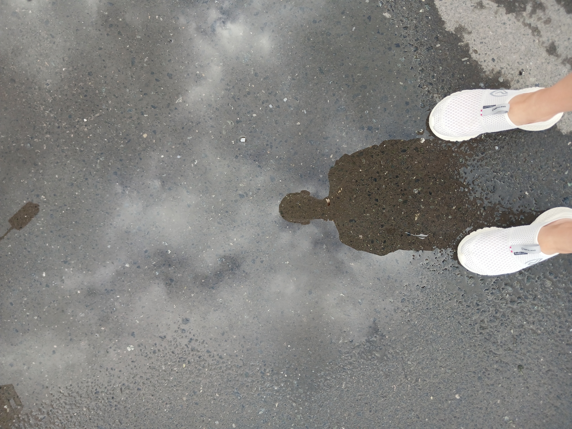 The image shows a reflection of a person's silhouette in a puddle on an asphalt surface. The person is wearing white sneakers. The reflection captures the head and shoulders of the person. The asphalt appears wet, suggesting it has rained recently. The sky is reflected in the puddle, showing cloudy conditions. The scene is outdoors, likely in a public space such as a street or sidewalk within the city of Yekaterinburg, Russia.