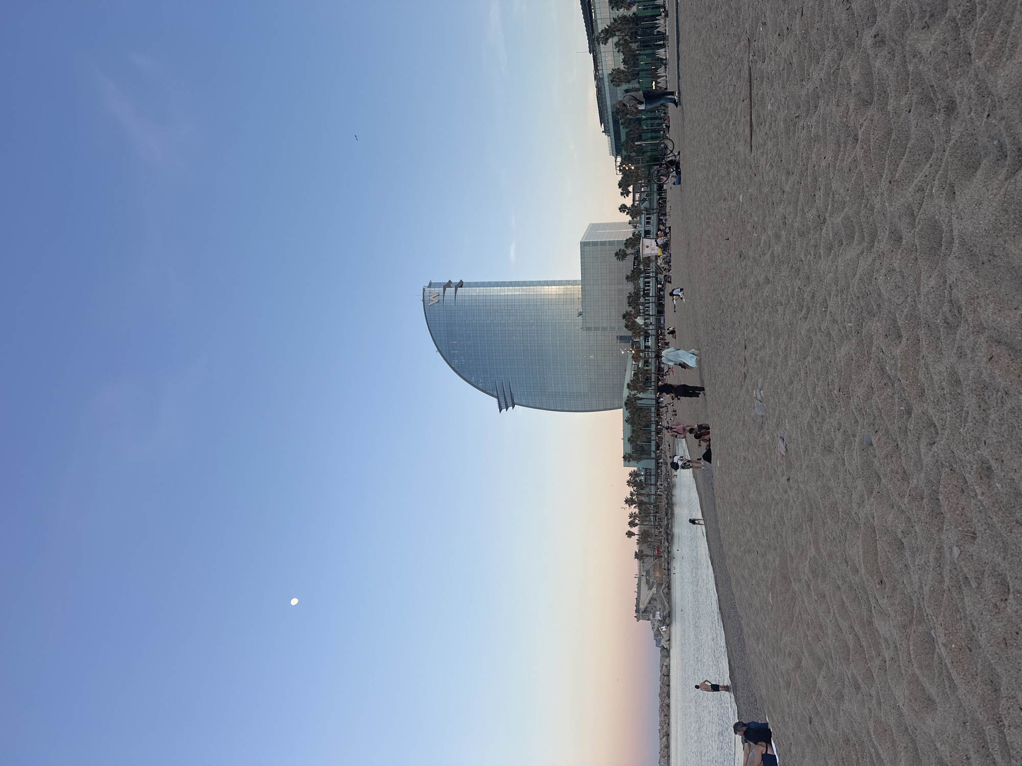 The image depicts a beach scene in Barcelona, Spain, at dusk. The foreground is dominated by a sandy beach with visible footprints and textures. In the mid-ground, the W Barcelona hotel, a distinctive glass-clad structure, stands out against the skyline. A line of palm trees and other buildings run parallel to the beach. People are visible along the beach, some walking and others appearing to be relaxing. The sky is a gradient of blue to orange, indicating the sunset. A small, circular object, likely the moon, is visible in the upper left portion of the image. There is also a bird flying in the sky.