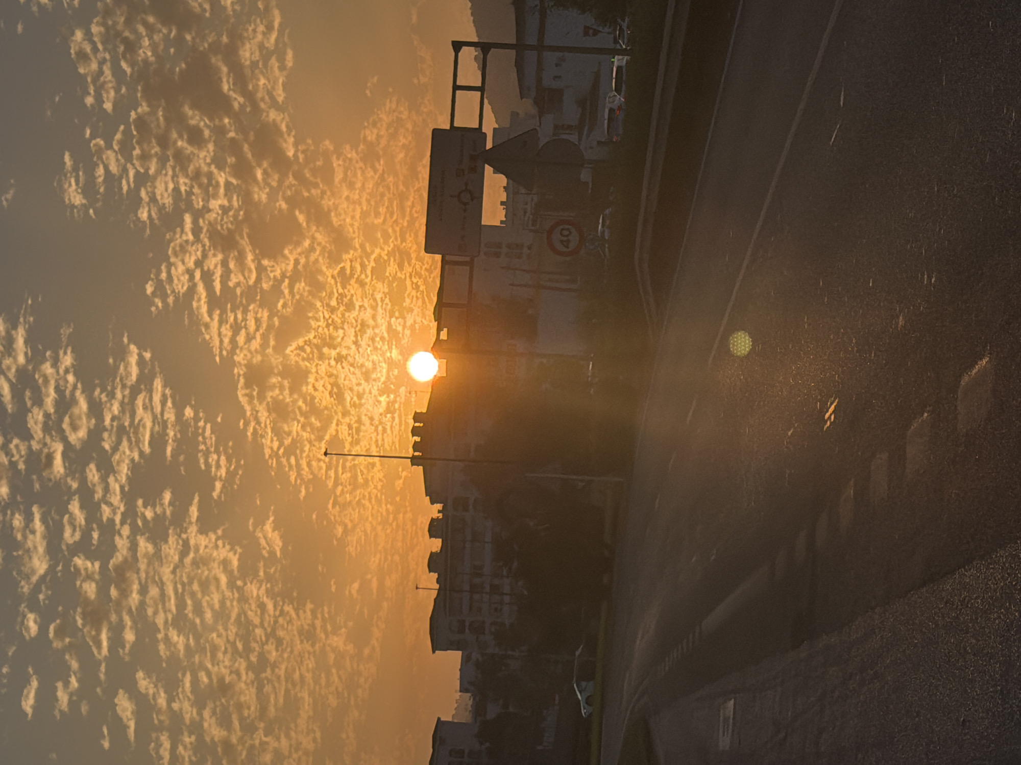 The image depicts a road in the city of Ibiza, Spain, during sunset. The sun, a bright orb, is positioned in the upper center of the frame, casting a warm orange glow across a sky filled with scattered clouds. In the background, silhouetted buildings with multiple stories and balconies are visible, indicative of an urban setting. A street light stands tall on the right side, its light diffused by the sunset. Road signs, including one with a circular "40" symbol, are present, suggesting the presence of traffic regulations. The road itself appears wet, reflecting the ambient light and indicating recent rain or humidity. Vehicles are sparsely visible on the road and in the background, suggesting moderate traffic. The overall atmosphere is serene, with the setting sun creating a dramatic and colorful sky.
