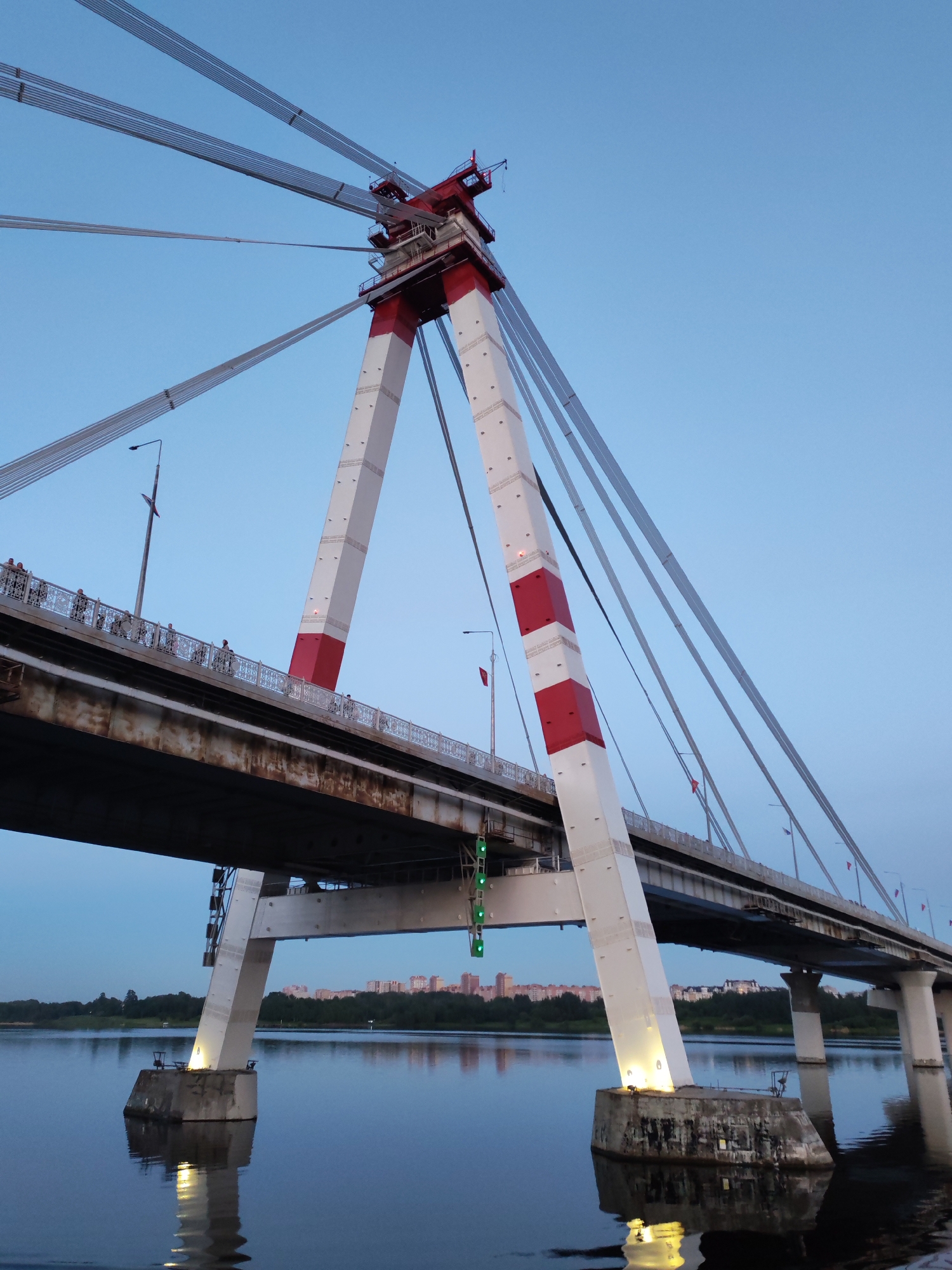 The image depicts a cable-stayed bridge at dusk. The bridge has a central pylon painted with alternating red and white sections. The pylon supports numerous steel cables that connect to the bridge deck. The bridge deck appears to be concrete and steel, with a pedestrian walkway along the side. People are visible walking on this walkway. Underneath the bridge deck, there are support structures that descend into the water. Buildings are visible in the distance. The water is calm, reflecting the bridge and the sky. The sky is a gradient of blue, suggesting it is either sunrise or sunset. There are flags flying along the bridge.