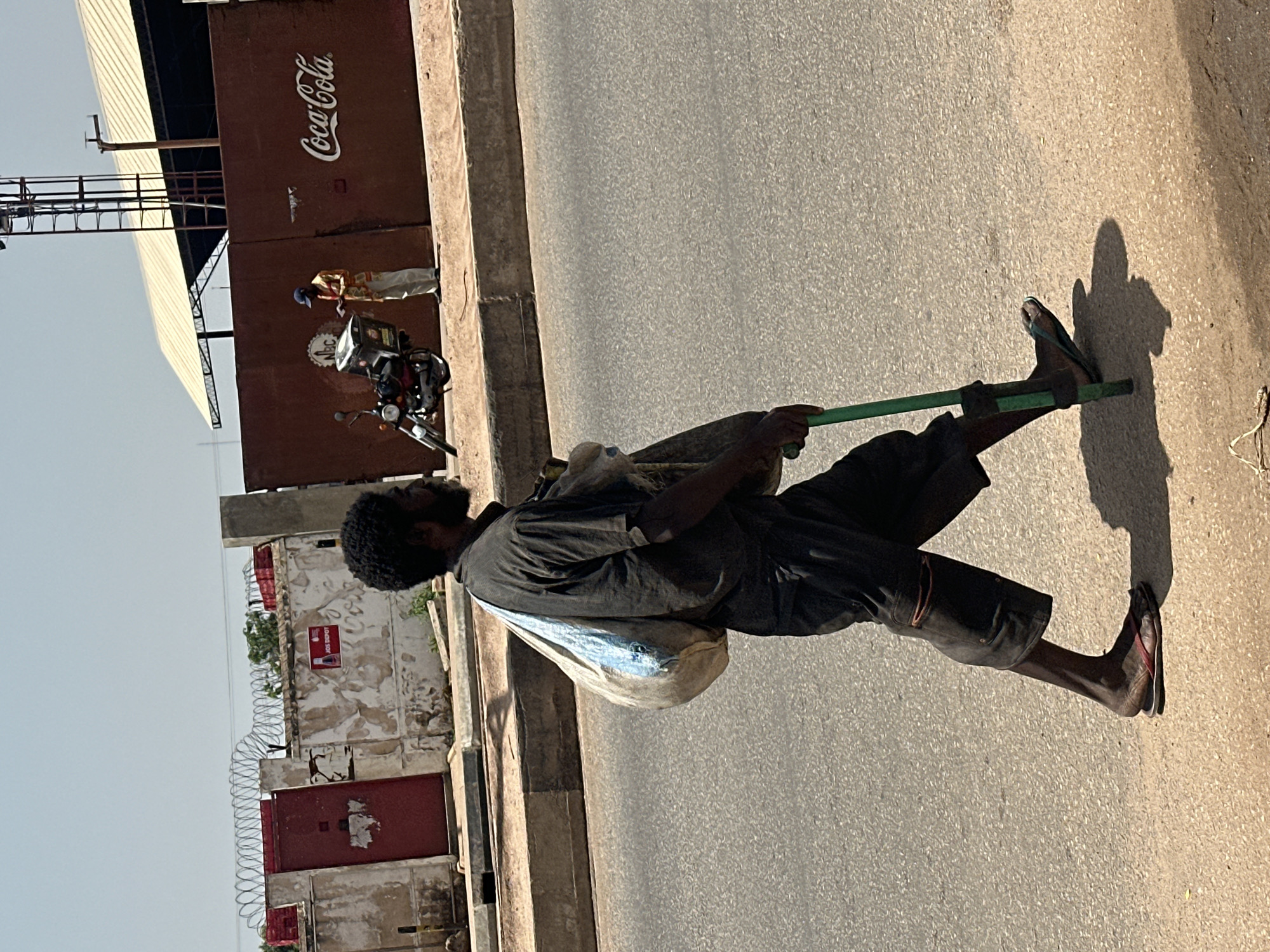 The image captures a street scene in Barakin Lerawi, Nigeria, on a bright, sunny day. In the foreground, a man with dark skin and curly hair walks on an asphalt road. He is carrying a large, light-colored bag over his left shoulder and a long, green pole in his right hand, which he appears to be using as a walking stick. He is wearing dark, loose-fitting clothing, including shorts that reach his knees, and dark flip-flops. His body is angled slightly away from the viewer, and his shadow is cast on the road behind him.
In the middle ground, a rusted brown wall forms a backdrop. On the wall, the Coca-Cola logo is prominently displayed in white lettering. To the left of the Coca-Cola logo, there is a circular emblem that reads "NBC". A motorcycle is parked in front of the wall, with a rider wearing a colorful shirt and a blue cap standing next to it, appearing to hold a piece of paper. Further to the left, a red gate is visible, topped with barbed wire.
The background shows a bright, hazy sky. Structures with slanted, light-colored roofs are visible in the distance. The environment appears to be an urban or industrial area, with a clear indication of commercial activity given the prominent branding. The road is clean, with a concrete curb separating it from the area behind the wall.