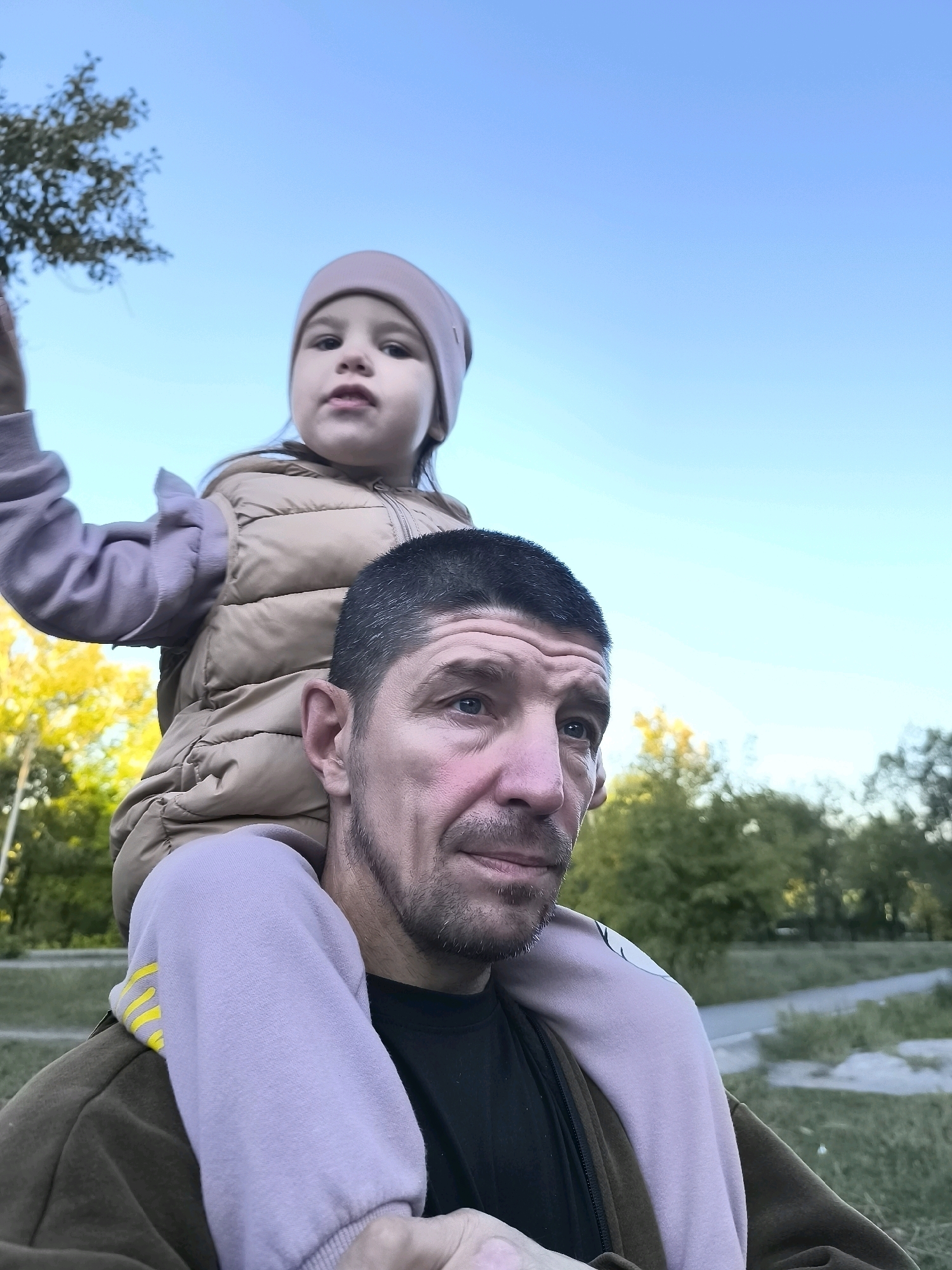 A close-up, low-angle shot captures a man with short, dark hair and stubble, looking to his right. He is carrying a young child on his shoulders, who is wearing a light pink beanie and a tan puffer vest over a purple hoodie with yellow stripes. The child's face is partially obscured by the man's head. In the background, trees with yellowing leaves and green foliage are visible under a clear blue sky. The scene appears to be an outdoor public space, possibly a park or walkway. The lighting suggests it is late afternoon or early evening. The image does not contain sufficient information to determine a specific location within the city of Qarağandı, Kazakhstan.
