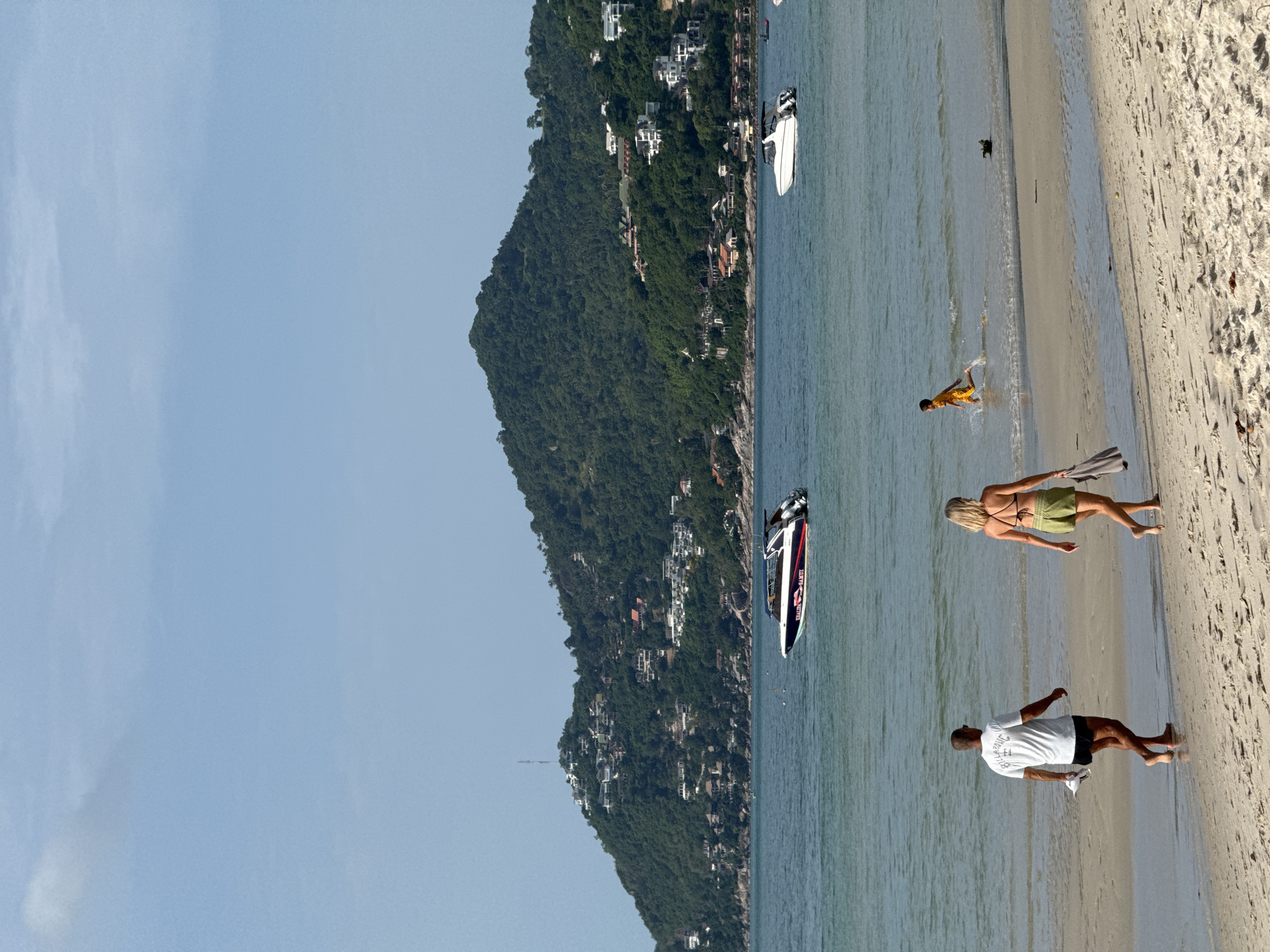 This media file depicts a sunny day at a beach in Ko Samui District, Thailand. The scene shows a tropical landscape with a lush green, tree-covered mountain rising from the coastline. Several white buildings are scattered across the hillside, indicating a developed area along the shore.
In the foreground, the sandy beach meets the clear, blue water. A few people are present. One person, wearing a yellow t-shirt and shorts, is standing in the shallow water, appearing to play or splash. Further down the beach, a woman in a bikini top and green shorts stands facing the water. Nearby, a man in a white t-shirt and black shorts is also on the sand, facing away from the camera.
Several boats are visible in the water. A white speed boat with red and black accents is positioned closer to the shore, while another white boat is further out. A small, dark object, possibly a dog, is also in the water near the shore. The clear sky suggests it is daytime, likely mid-morning or afternoon, with good weather. The overall atmosphere appears relaxed and leisurely, typical of a beach vacation. The text "TOMMY NUNAN" is visible on the side of the speed boat.