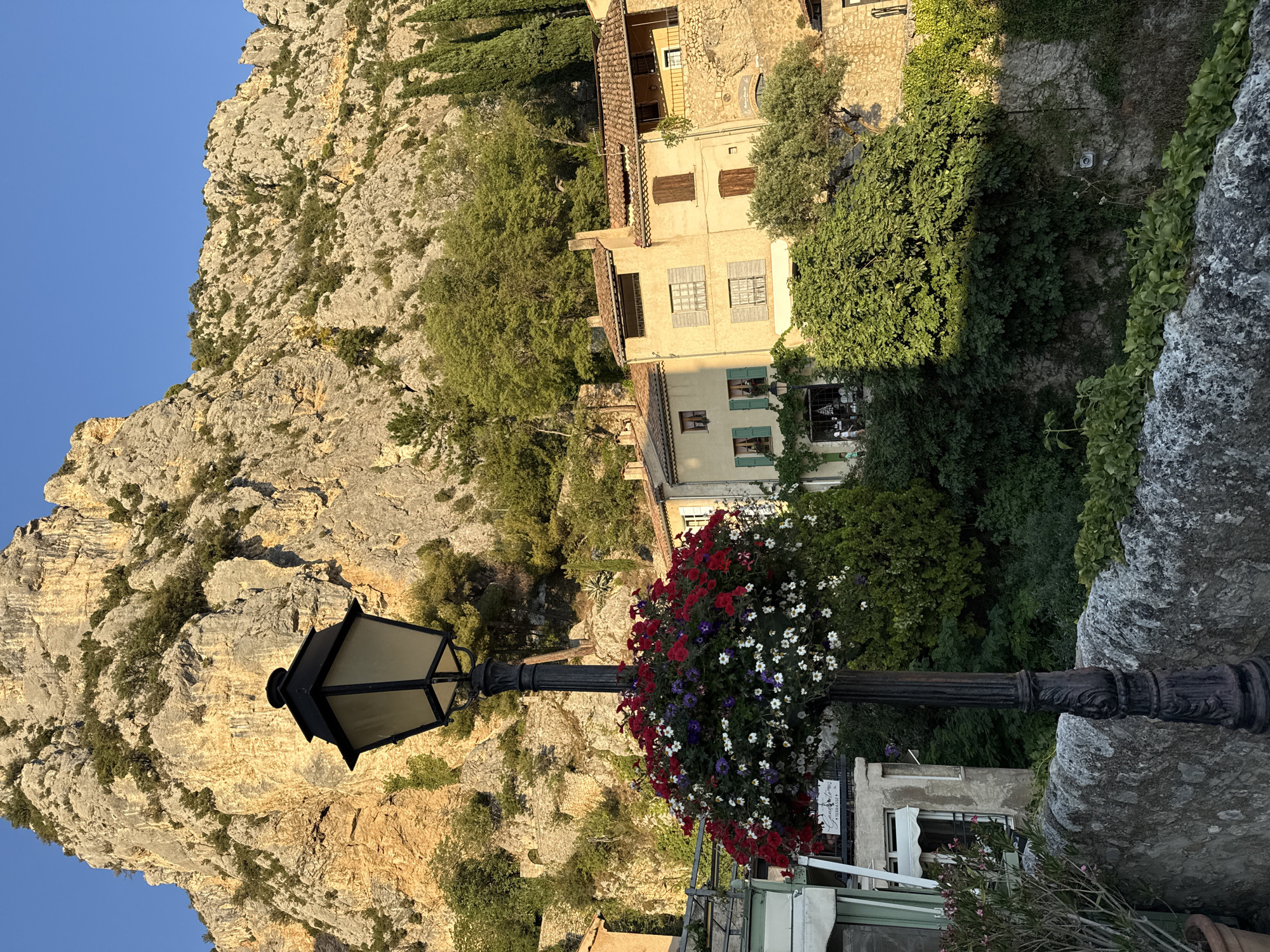 A lamppost decorated with a hanging basket of red, white, and purple flowers stands in front of a rocky mountain and a village in Moustiers-Sainte-Marie, France. The lamppost is black and ornate, with a square lantern at the top. The hanging basket is full of vibrant flowers, creating a beautiful contrast with the natural landscape. Behind the lamppost, the village buildings are made of stone and stucco, with red tile roofs. The mountain is rugged and steep, with trees and vegetation growing on its slopes. The sky is clear and blue, and the sun is shining, casting a warm glow on the scene.