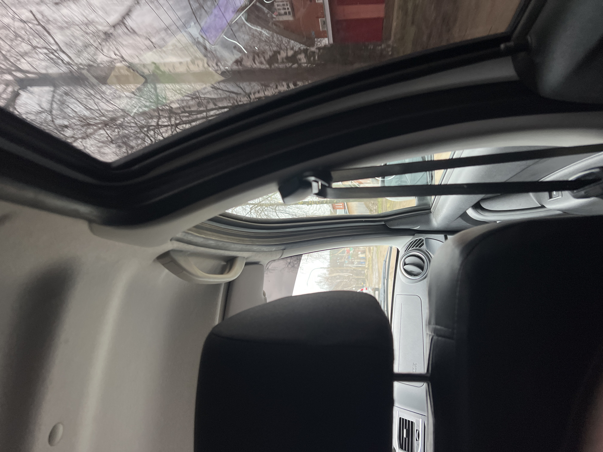 The media file presents an interior view of a vehicle, captured from a rearward perspective looking towards the front and upwards through a sunroof. The image is rotated, showing the roof and the sky on the left.
The dominant feature is an open sunroof, revealing a view of an overcast sky and numerous bare tree branches, suggesting a cold season like winter or early spring. Reflections on the sunroof glass also show parts of a red-brick building.
Inside the vehicle, the light-colored headliner and a gray grab handle are visible on the left side. The black fabric headrest of the front passenger seat occupies the lower right portion of the frame. To the right of the seat, a circular air vent is prominent on the dashboard. Further forward and lower, a rectangular air vent is also visible. Faint text reading "SRS AIRBAG" is discernible on the passenger side dashboard, indicating an airbag location.
Through the front windshield, more bare trees and an indistinct background suggesting a residential street can be seen. The vehicle appears stationary. There are no people visible, and no specific activity is taking place. The overall impression is of a quiet, possibly overcast day in a location like Chaikovsky, Russia, as indicated by the context.