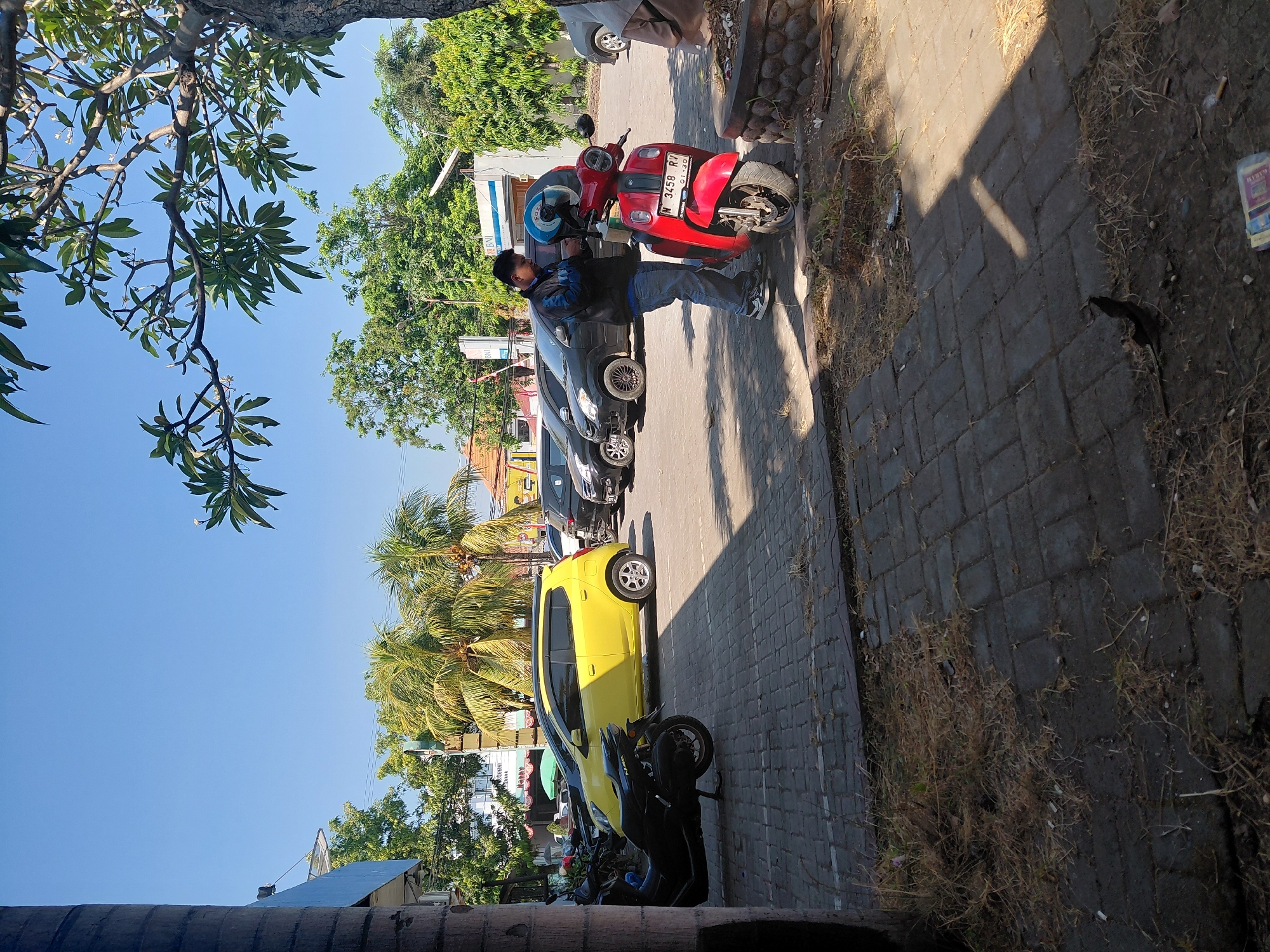 A man in a blue and black jacket and blue jeans is standing next to a red scooter in a parking lot under a clear blue sky. The scooter has a license plate that reads "N 3458 RV 01-30". Behind the scooter, a yellow car is parked next to several dark-colored cars. Palm trees and other green trees are visible in the background, along with a building that appears to be a bank with a "BNI" logo. The ground is paved with interlocking bricks, and dry grass and leaves are scattered on the ground. The image appears to be taken during the day. This scene is located in Semampir, Indonesia.
