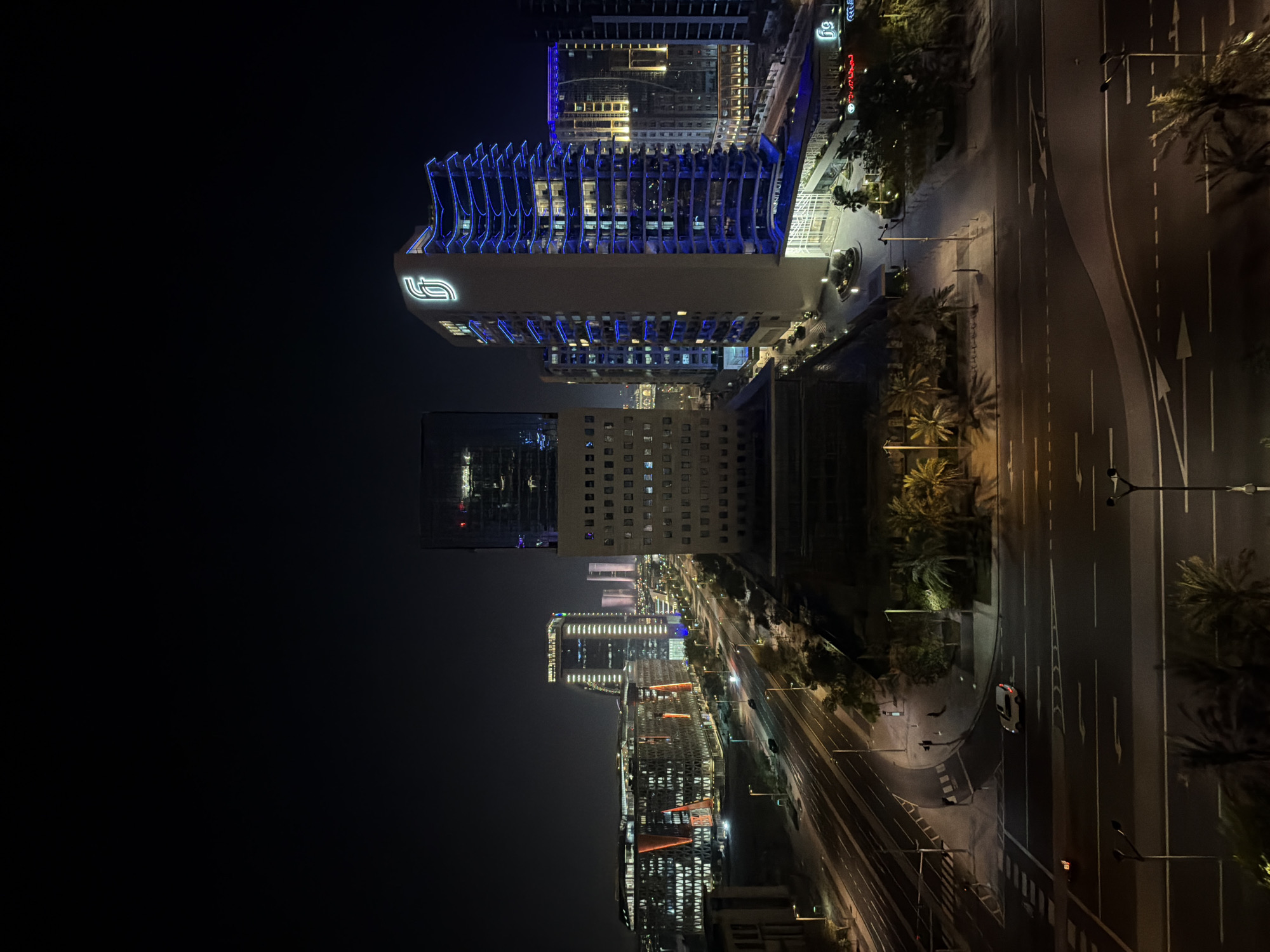 The image shows a city scene at night with modern architecture and street lights. In the foreground, there are multiple lanes of a road with traffic signals and street lamps. A white car is visible on the road. Palm trees are scattered along the sides of the road. In the midground and background, several tall buildings are illuminated with lights, some with blue and orange accents. One prominent building has a glowing logo at the top. The sky is dark, indicating nighttime. The scene appears to be a business or commercial district within a city.