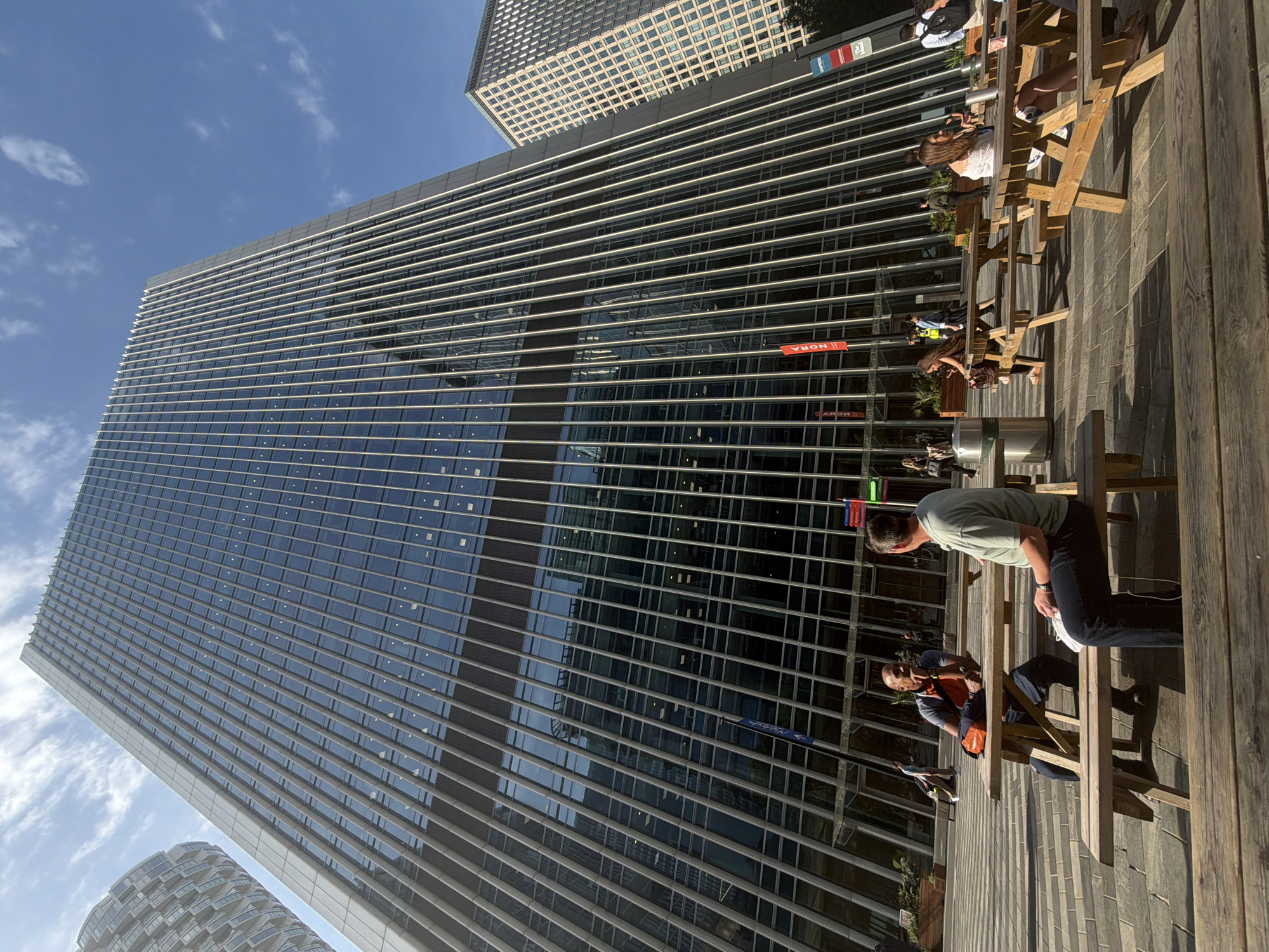 The image shows an outdoor scene in the city of London, United Kingdom. A modern, towering skyscraper with a grid-like facade dominates the left side of the frame. Adjacent to this is another skyscraper with a similar design.
To the right, a wooden deck is visible, furnished with wooden picnic benches and tables. Several people are seated at these benches, some engaged in conversation and others seemingly relaxing. A man in a light green shirt is standing beside one of the benches, leaning slightly forward. Other people can be seen further along the deck, with varying poses and actions.
The sky is blue with some scattered clouds, suggesting a clear day. The overall impression is of a public space in a city environment, possibly a patio or outdoor seating area for a restaurant or office building.