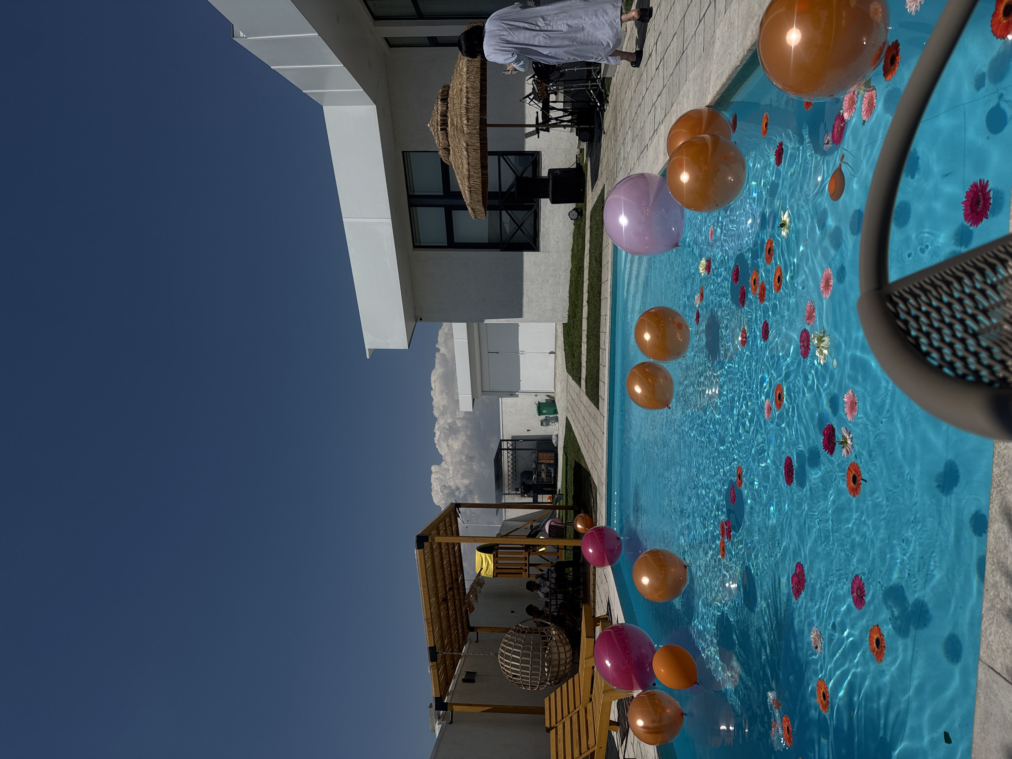 The image depicts an outdoor swimming pool area, likely in Proshyan, Armenia, on a bright, clear day. The pool itself is filled with turquoise water and decorated with numerous floating flowers in various colors, including pink, orange, and white. Several large, inflated balloons in shades of pink, orange, and iridescent purple are also floating on or near the surface of the water.
In the background, to the left, a wooden pergola with a thatched roof covers a seating area where a group of people are gathered around a table. Adjacent to this is a children's play structure with a yellow slide. Further to the right, a modern white building with dark-framed windows is visible. Near the building, a woman in a light blue, long-sleeved dress is standing on a paved area, holding a phone and looking towards the pool. A thatched umbrella stands nearby, and behind it, musical equipment, including speakers and a drum set, is set up. The ground around the pool and building is paved, with patches of green grass. The overall scene suggests a festive gathering or party.