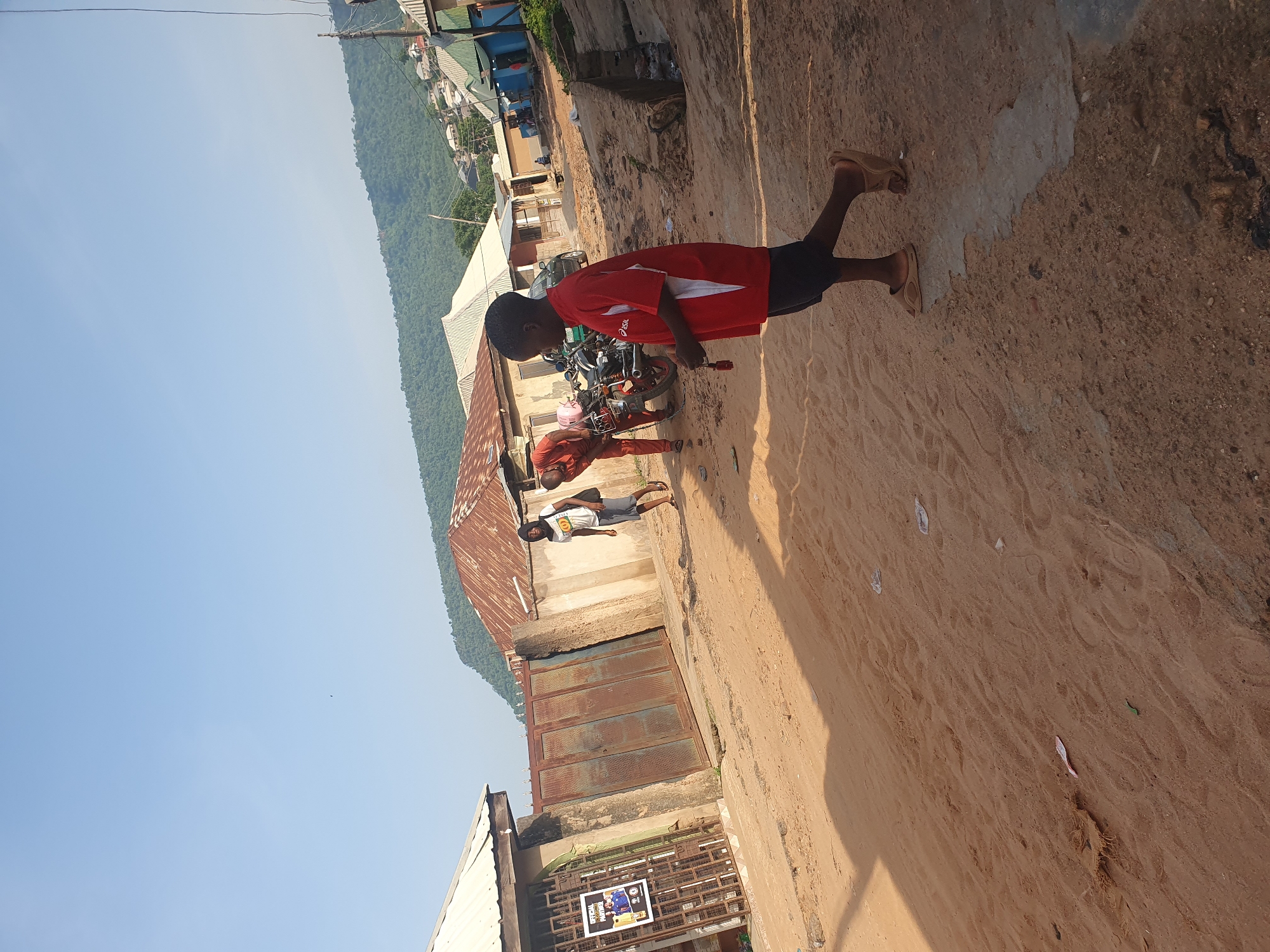 The image depicts a sandy street scene in Lokoja, Nigeria, with a mountainous backdrop. In the foreground, a young boy in a red t-shirt and black shorts walks away from the camera, wearing beige sandals. To his left, a man in an orange jumpsuit bends over a motorcycle. Another man wearing a white t-shirt and grey shorts stands nearby. Buildings with corrugated iron roofs line the street, some appearing weathered. A large, rusty metal gate is visible on the left side of the street, with a poster attached to a nearby metal fence. The ground is uneven and sandy, with some scattered debris and footprints. The sky is clear and blue.