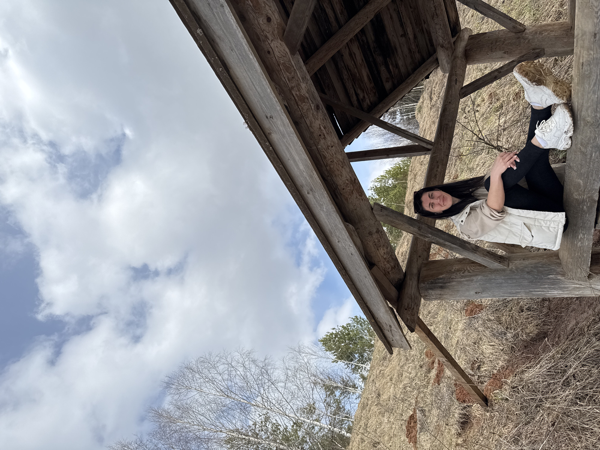 A woman is sitting in a wooden structure in Вассята, Russia. The wooden structure appears to be a small, open-sided shelter or gazebo, likely located in a natural environment, as evidenced by the trees and foliage visible in the background. The ground outside the shelter appears to be covered with dry vegetation. The sky is partially covered with clouds.
