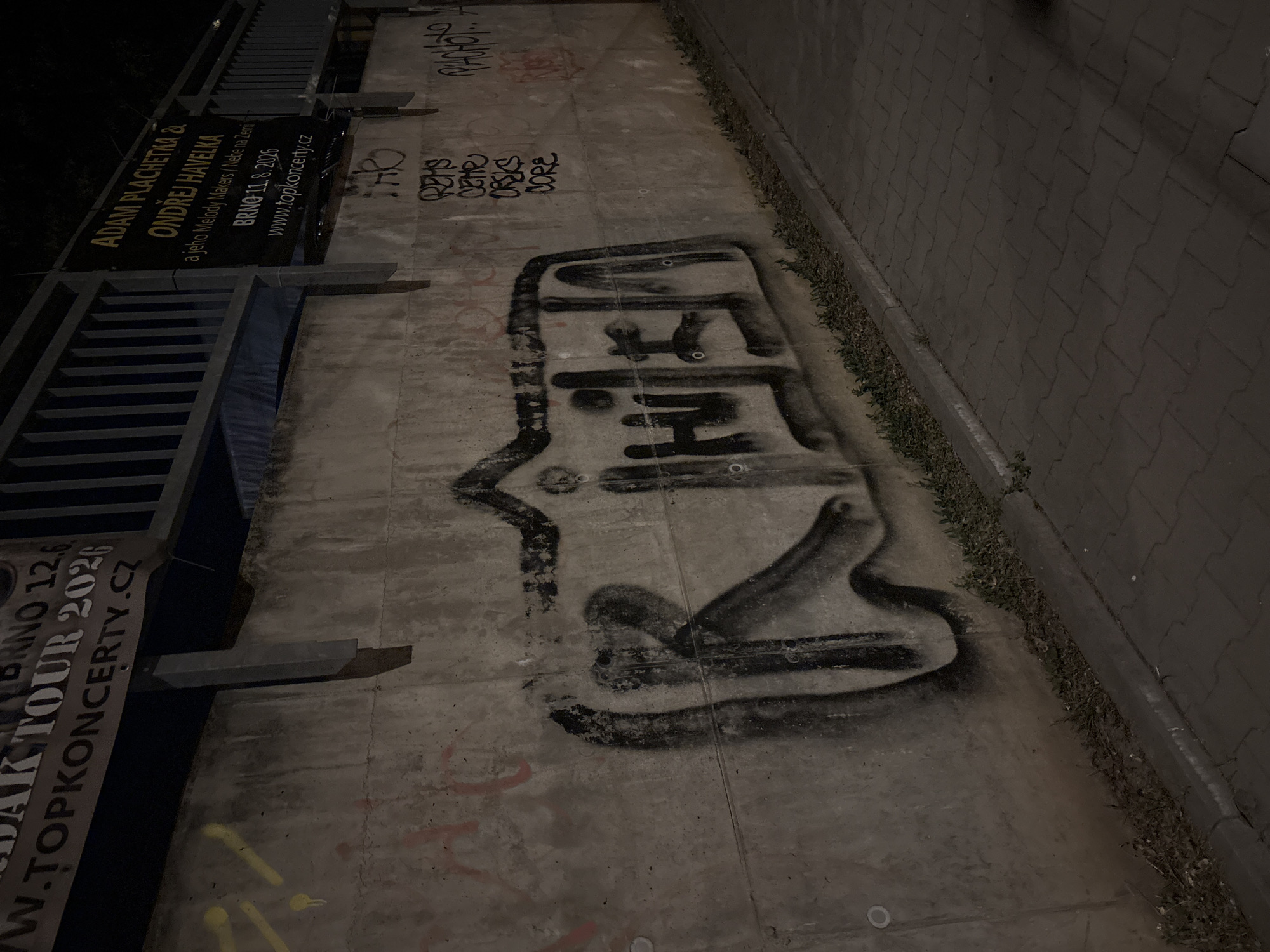 The image is an outdoor nighttime photograph of a concrete wall with graffiti. The wall is likely part of an underpass or bridge structure, as indicated by the railing and signage visible at the top of the frame. The graffiti on the wall is primarily spray paint. A large, stylized tag in black spray paint dominates the center of the wall, with other tags and markings in black and red scattered across the surface. To the right of the main tag, there are smaller, vertically aligned tags in black spray paint. The ground in front of the wall is paved with interlocking bricks and a strip of grass and dirt is visible between the pavement and the wall. The overall lighting is dim, suggesting it is nighttime, with artificial light sources illuminating the scene. A banner with text in English and Czech is attached to the railing above the wall.