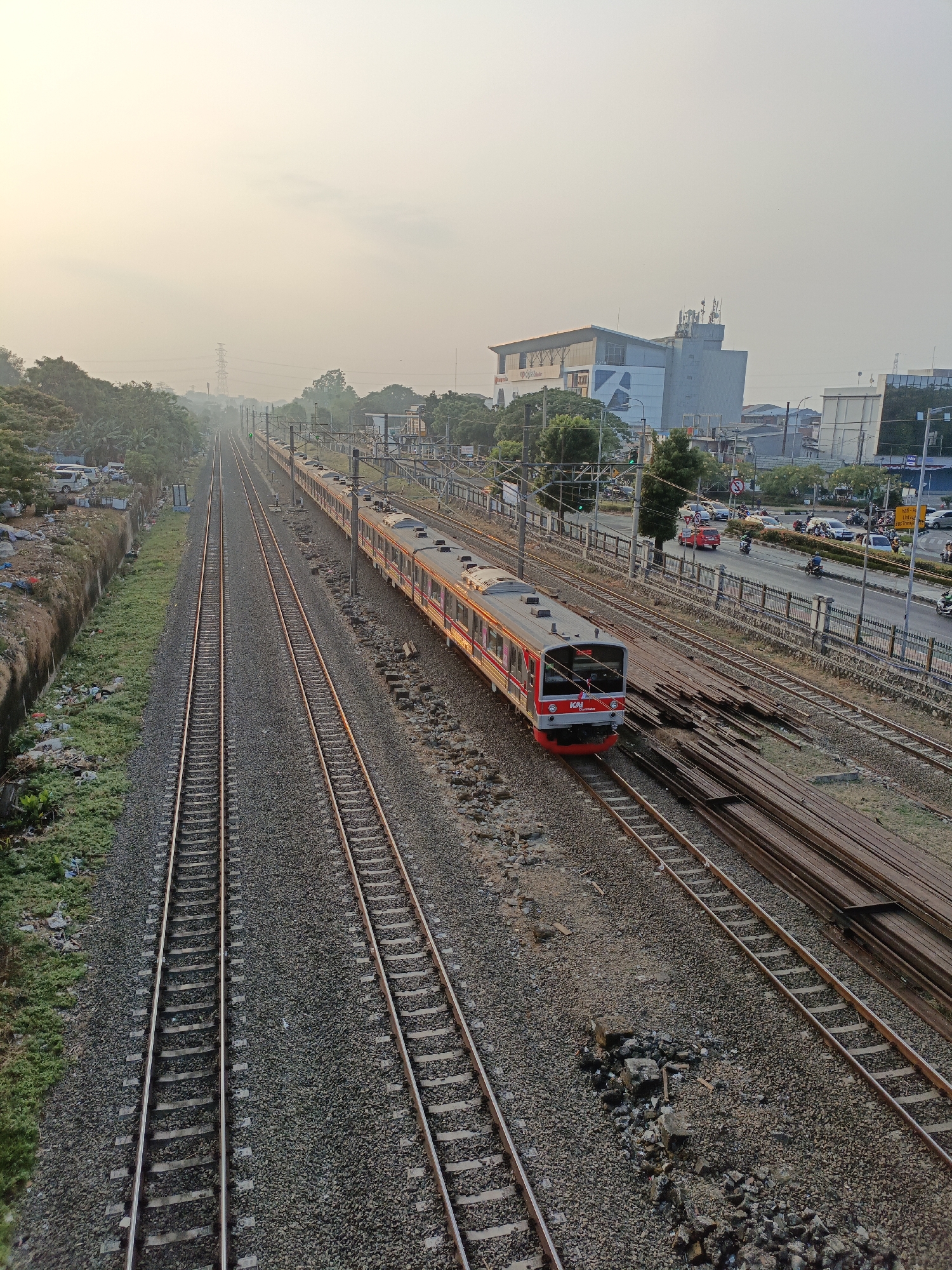 The image captures a train on a railway track in East Jakarta, Indonesia, during what appears to be dawn or dusk, indicated by the hazy, bright sky. The train, predominantly red and white, is positioned on the right set of tracks, moving away from the viewer. The railway consists of multiple parallel tracks, with gravel ballast. To the left of the tracks, a slope is covered with green grass and some debris, with parked cars visible at the top. Beyond the tracks, a road runs parallel, with cars, motorcycles, and pedestrians present. Several buildings, including a multi-story structure with large windows and antennas on its roof, are visible in the background, suggesting an urban environment. Overhead power lines and transmission towers are also present in the distance. The overall scene depicts a functional urban landscape with active transportation infrastructure.