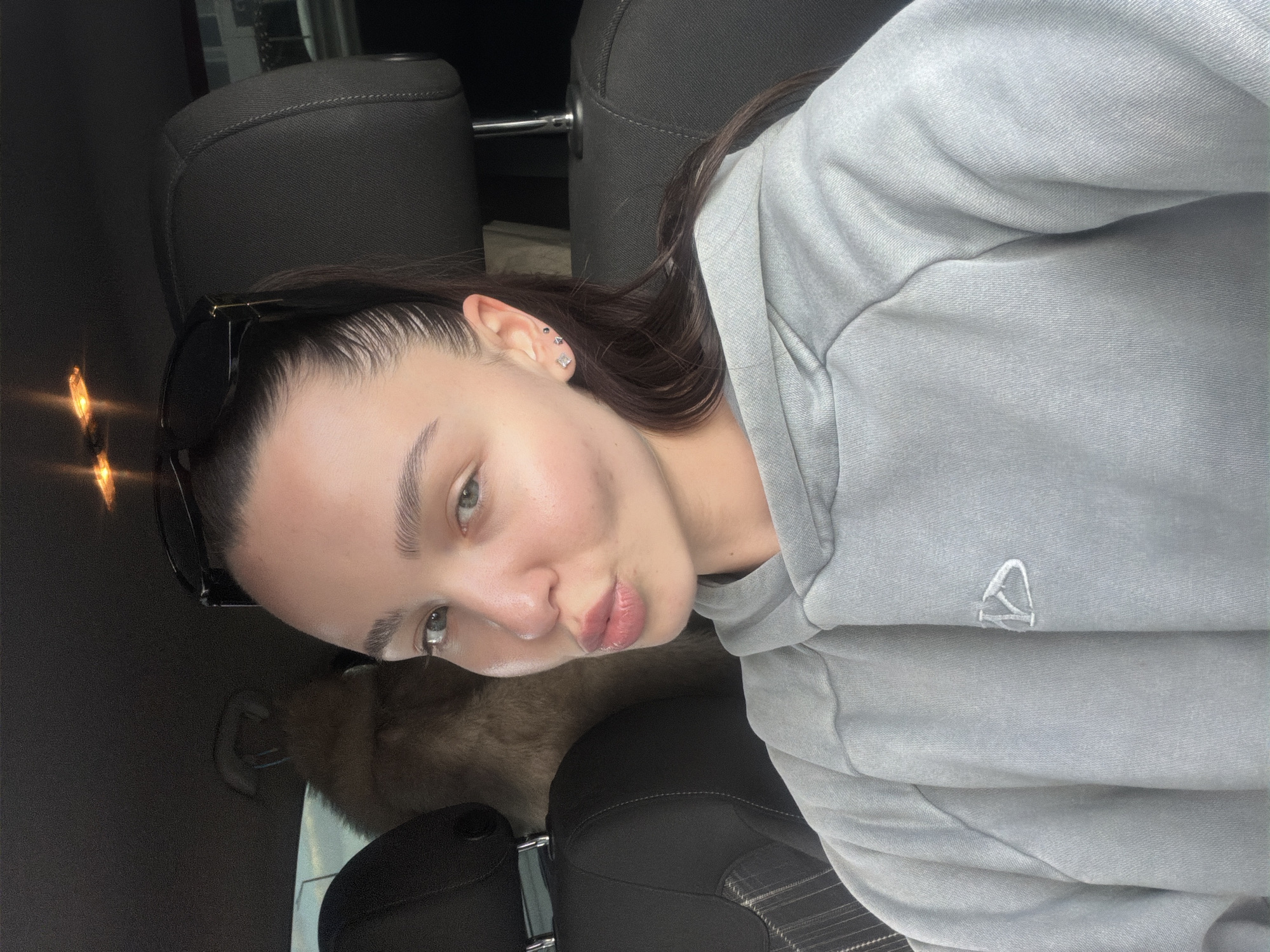 A young woman is featured in a close-up selfie taken inside a car, with the image rotated 90 degrees clockwise. She has long, dark hair pulled back, with black sunglasses resting on top of her head. Her face is turned slightly towards the camera, and she is making a playful "pout" with her full, slightly tinted lips. Her light-colored eyes look directly at the viewer, framed by prominent, well-defined eyebrows. Several small, silver stud piercings, including a square and a triangular one, are visible in her right ear.
She is wearing a light gray hooded sweatshirt with a small, white, stylized logo resembling an abstract letter 'A' or an upward-pointing arrow on the chest. The background shows the dark interior of a car, including the fabric of a headrest behind her. A fuzzy, brown object, possibly a pet or a fur-covered item, is partially visible on the seat below her face. Warm, glowing light reflections, likely from external sources or interior car lights, appear on the left side of the frame. The overall mood is casual and relaxed. The location context is Sofia, Bulgaria.