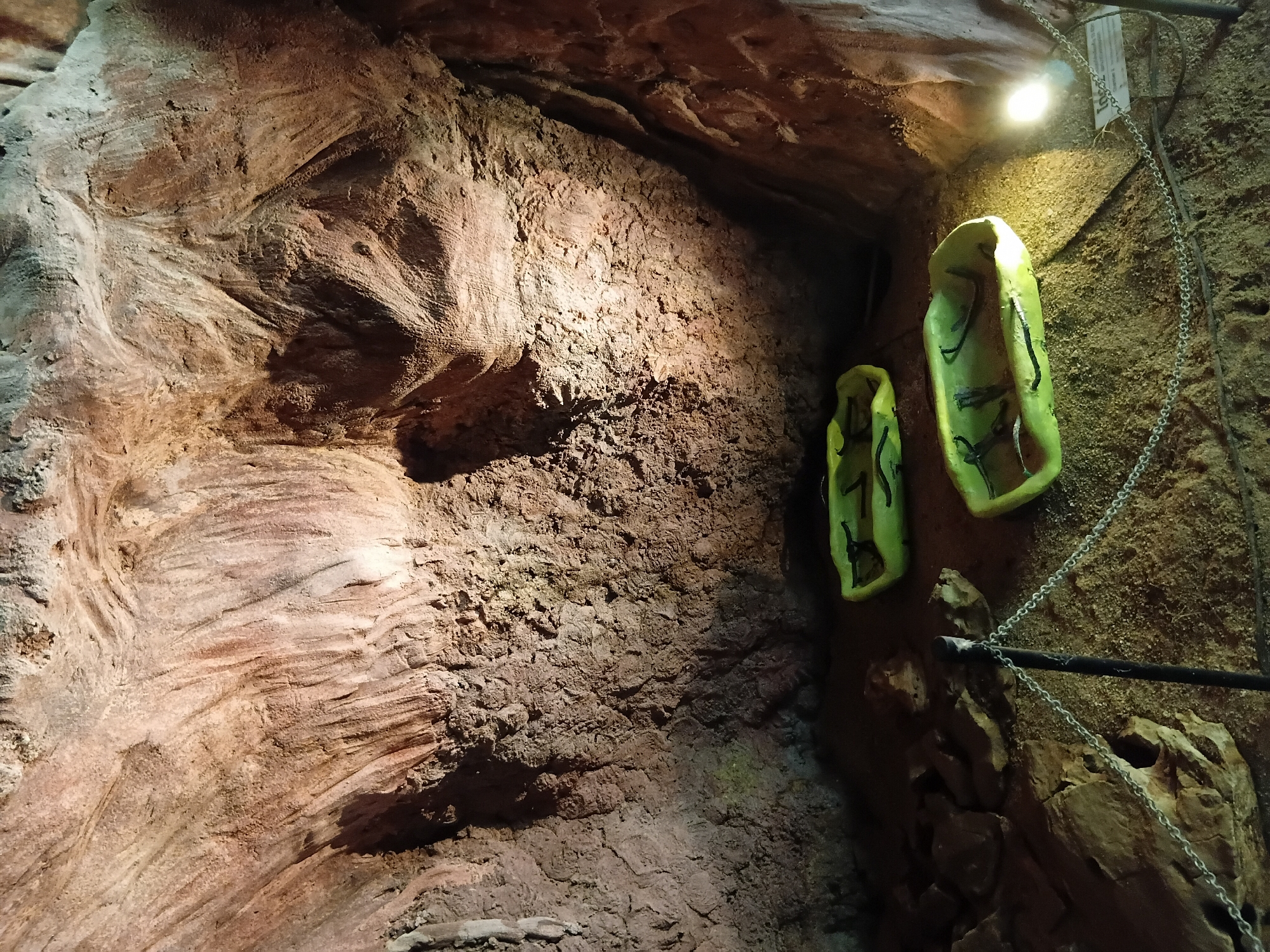 This image shows an exhibit inside a cave in Mae Sai District, Thailand. The cave walls are textured and appear to be made of rock. Two bright green, boat-shaped displays are on the sandy cave floor, each containing what appear to be black, elongated objects resembling bones or fossils. The displays are separated by a short distance. A metal chain fence with a black post in the foreground separates the viewer from the exhibit. A bright light source illuminates the right side of the scene, casting a glow on the cave wall and one of the displays. A small informational sign is visible near the light source. The overall impression is of a museum exhibit set within a natural cave environment.