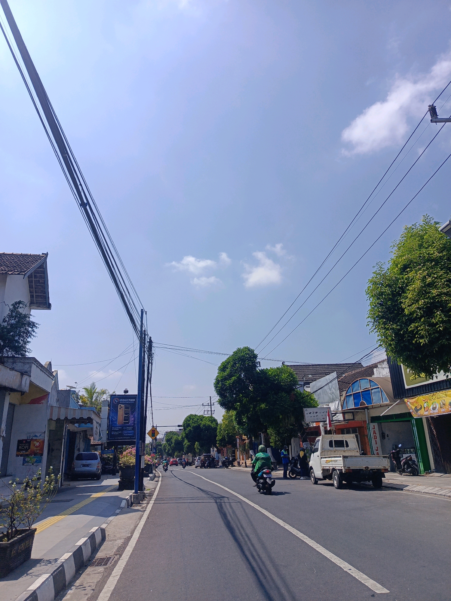 The image depicts a sunny street scene in Magetan, Indonesia, likely during daytime. The perspective is from a street-level view looking down a paved road lined with buildings and trees.
In the foreground on the left, a sidewalk with a textured yellow strip runs alongside the road. Several potted plants are visible on the sidewalk. The road itself is asphalt with a white dividing line.
Several vehicles are present on the road and parked along the sides. A white pickup truck is parked on the right side of the road. A person on a green motorcycle is riding in the middle of the road, heading away from the viewer. Other vehicles, including cars and motorcycles, are scattered further down the road.
Buildings on both sides of the street house various establishments. On the left, a building with a tiled roof is visible, as well as a storefront with signage, including what appears to be an advertisement for "MENDANY PHONE." On the right, several storefronts are present, one with a green awning and another with colorful signage featuring food items. Trees with lush green foliage line the street, providing shade.
Power lines crisscross the sky, supported by poles. The sky is clear blue with a few scattered white clouds. The overall impression is a typical bustling street in a provincial Indonesian town.